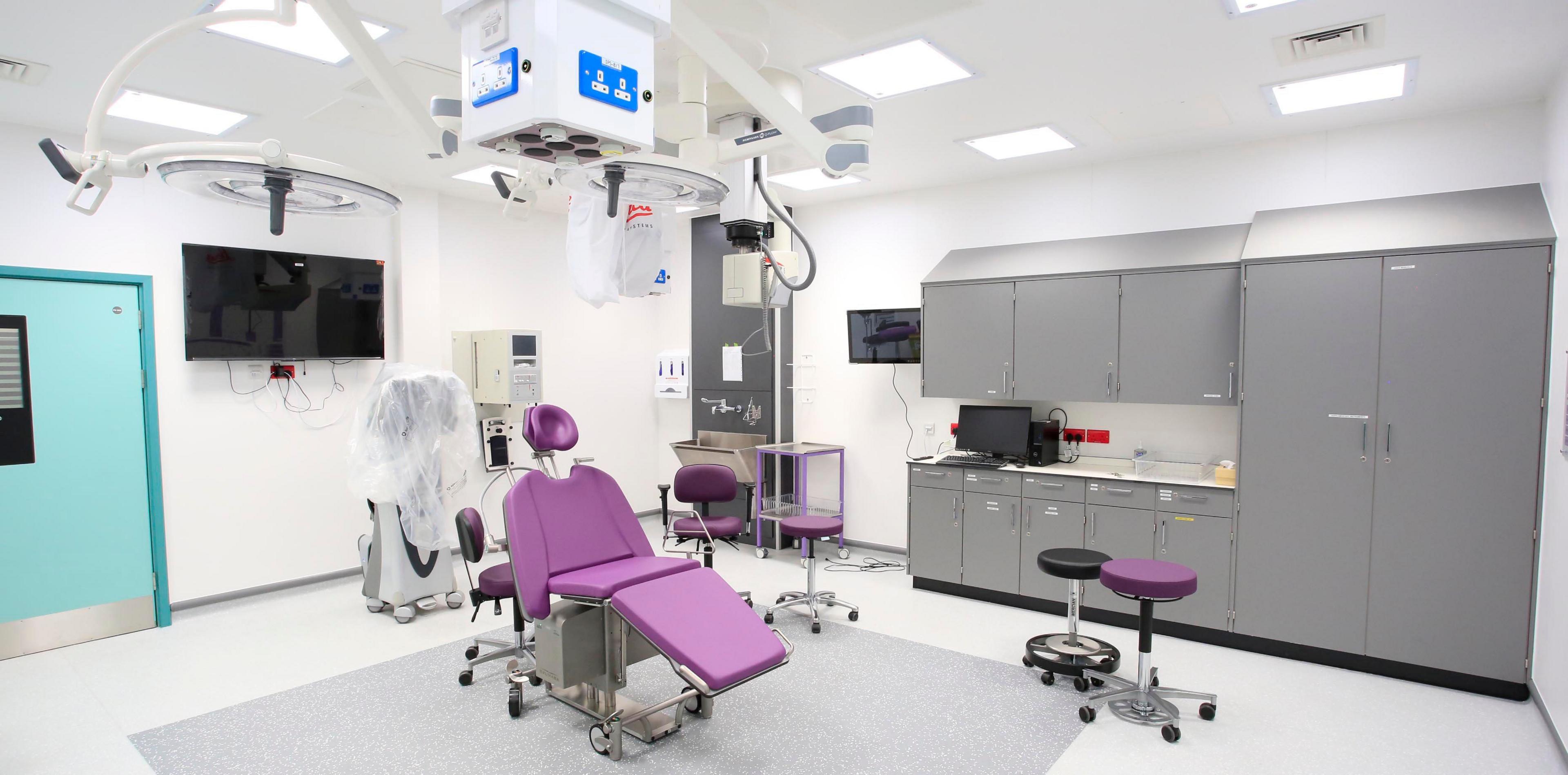 A purple opticians chair in a surgical suite. Two stools, one purple and one black are to the right of the chair 