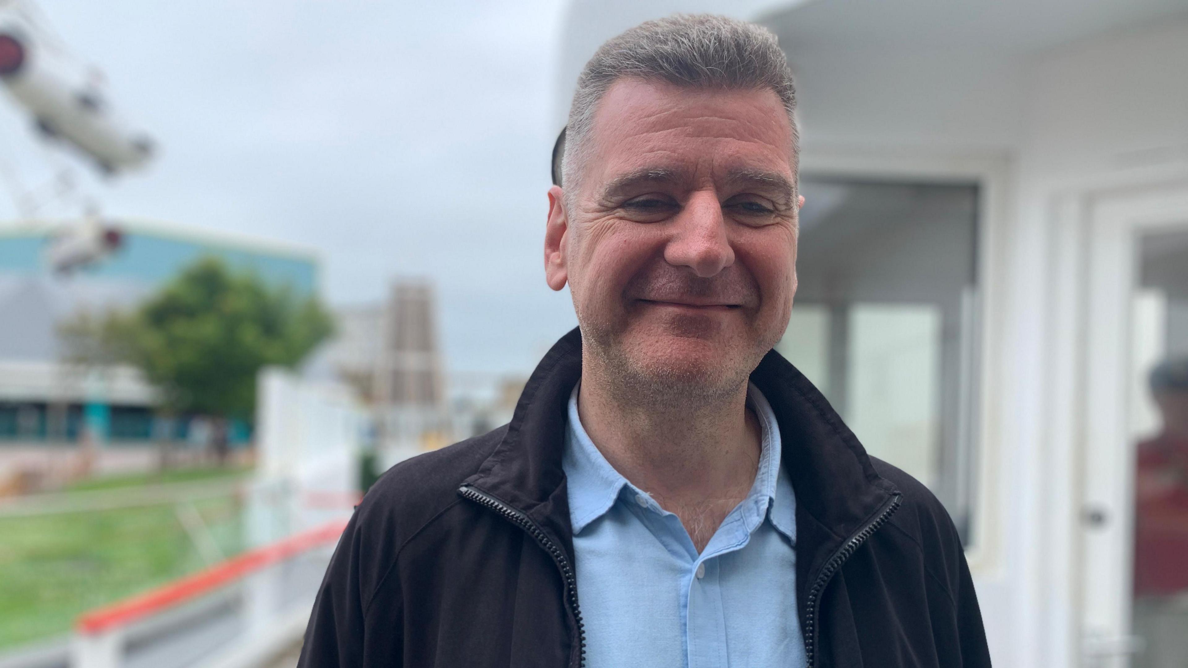 A middle-aged man stands outside wearing a light blue button-up shirt and and a black jumper. He is smiling.
