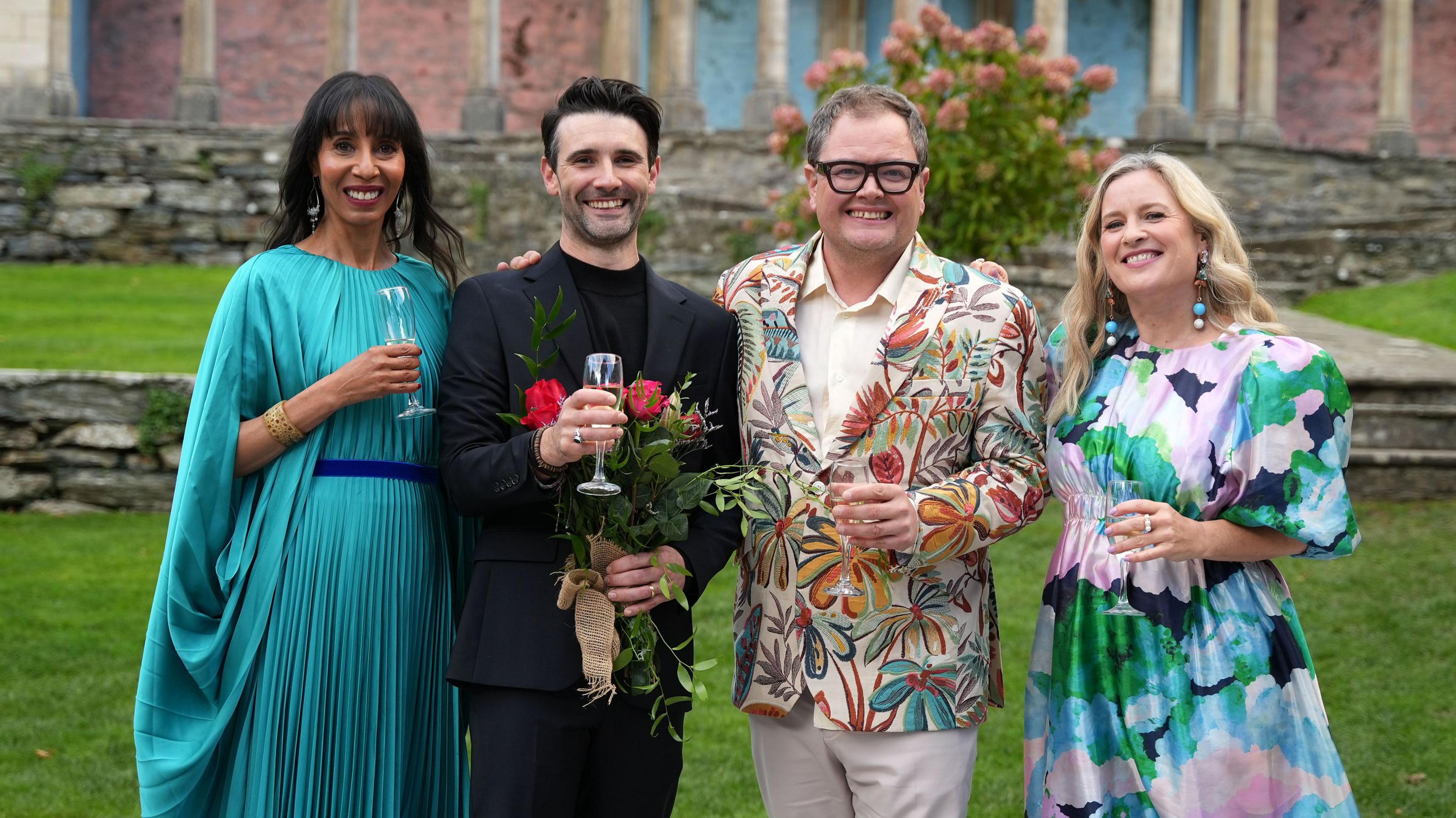 John Cooper wears a black suit and stands holding flowers and a glass of champagne next to Alan Carr. Elegantly-dressed women stand on either side of them, also each holding glasses of champagne.
