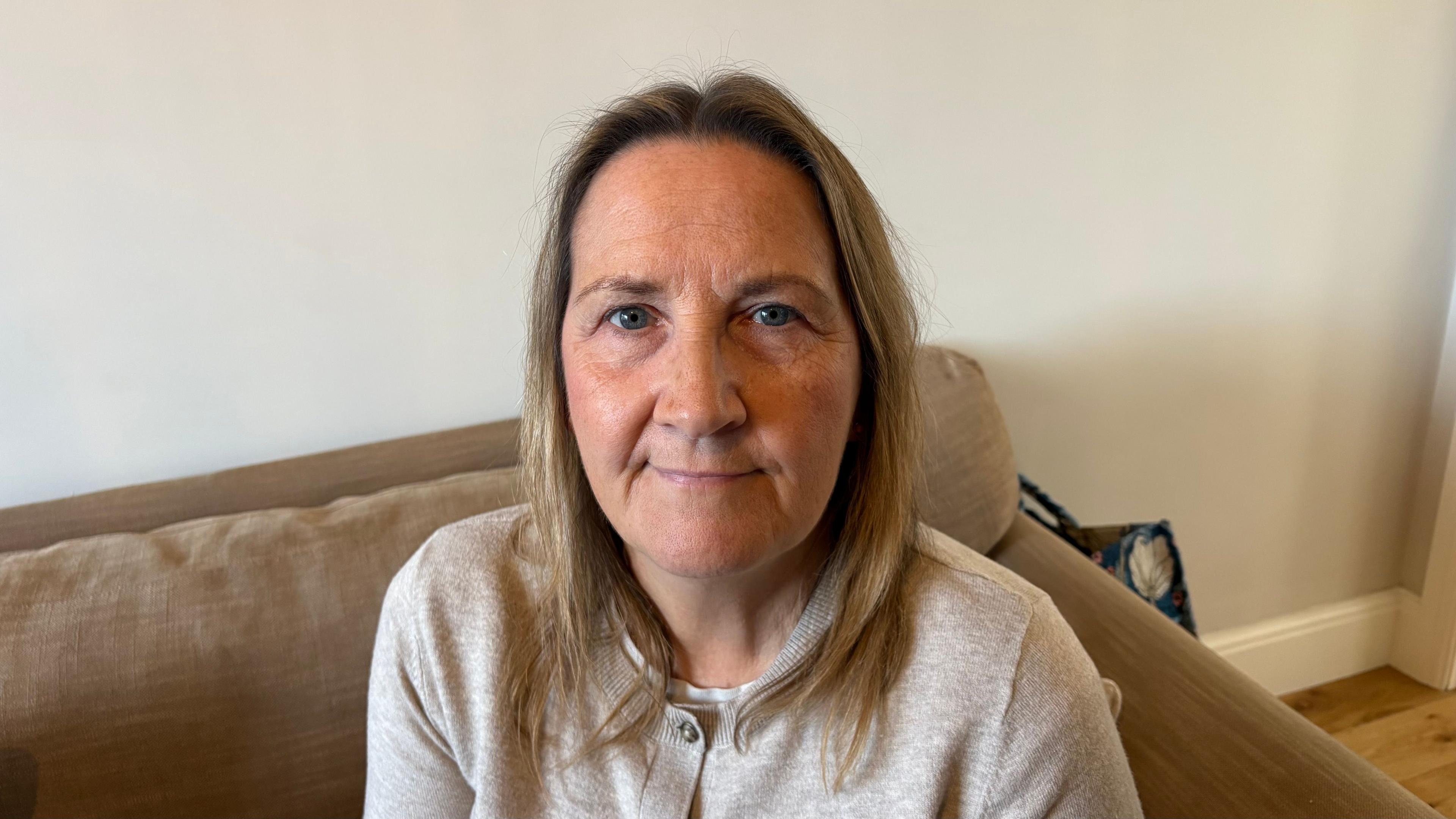 Tracey Hanson with long light brown hair, wearing a beige top and sitting on a brown sofa against a white wall