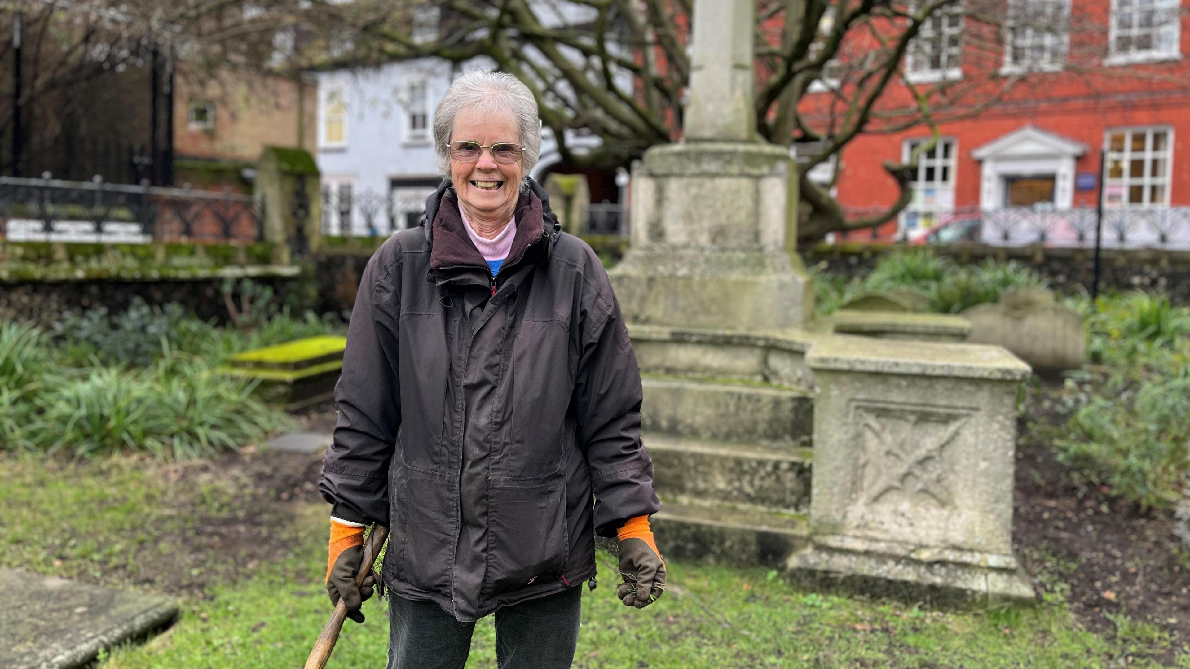 June Molloy standing with a garden trowel in front of a stone memorial in the church's grounds, smiling at the camera. She has white hair, glasses and is wearing a wax jacket over various layers of clothing.