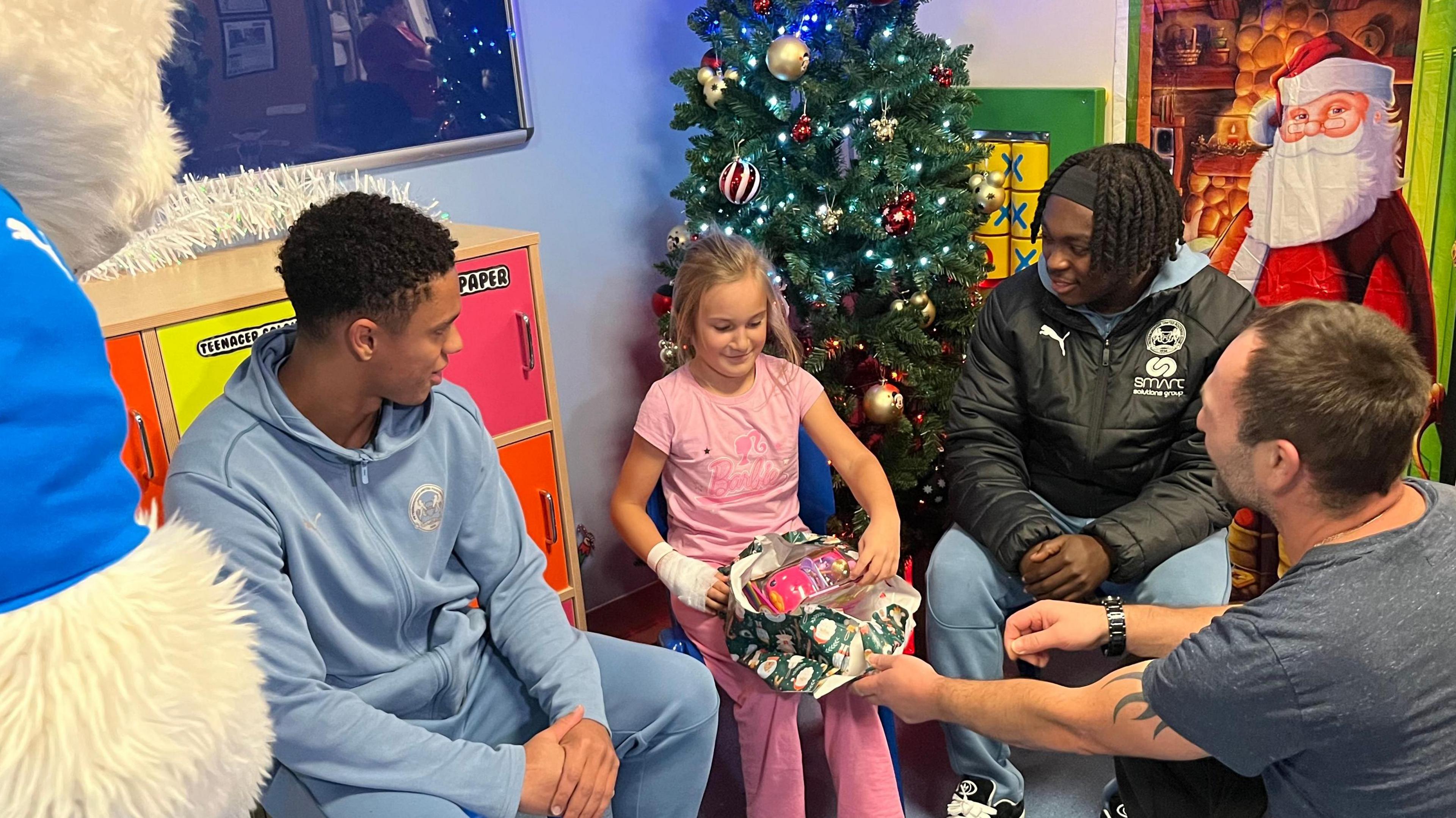 Posh players, wearing light blue branded tracksuits, are sitting in the hospital and watching Leja un-wrap a present. Leja is smiling and wears a pink barbie top and matching pants. Her toy looks like a pink car with a doll inside. 