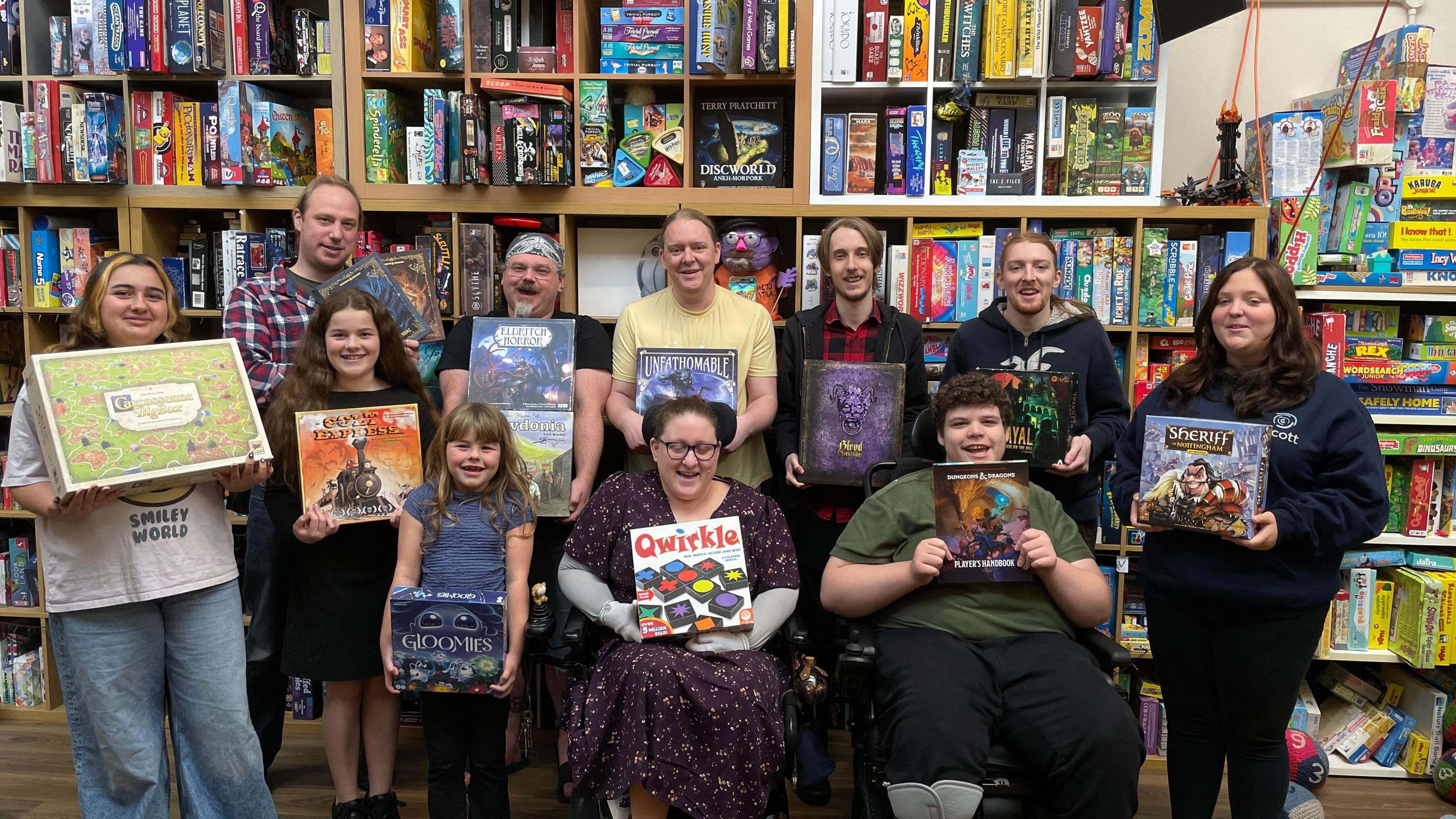 A group of people stand holding board games, smiling. Behind them is a wall of shelves filled with board games.