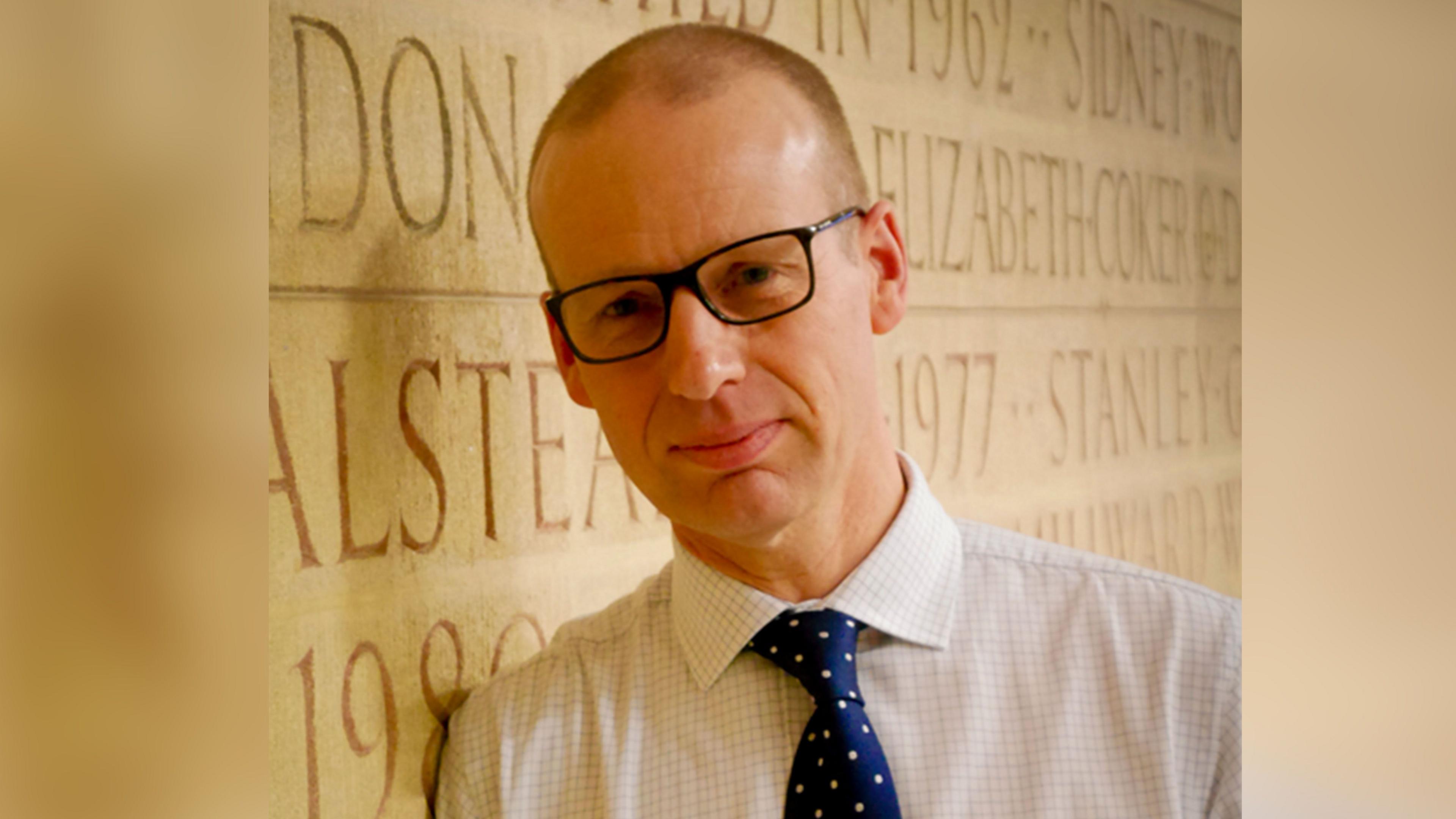 Richard is looking into the camera wearing black glasses a blue tie and white shirt.