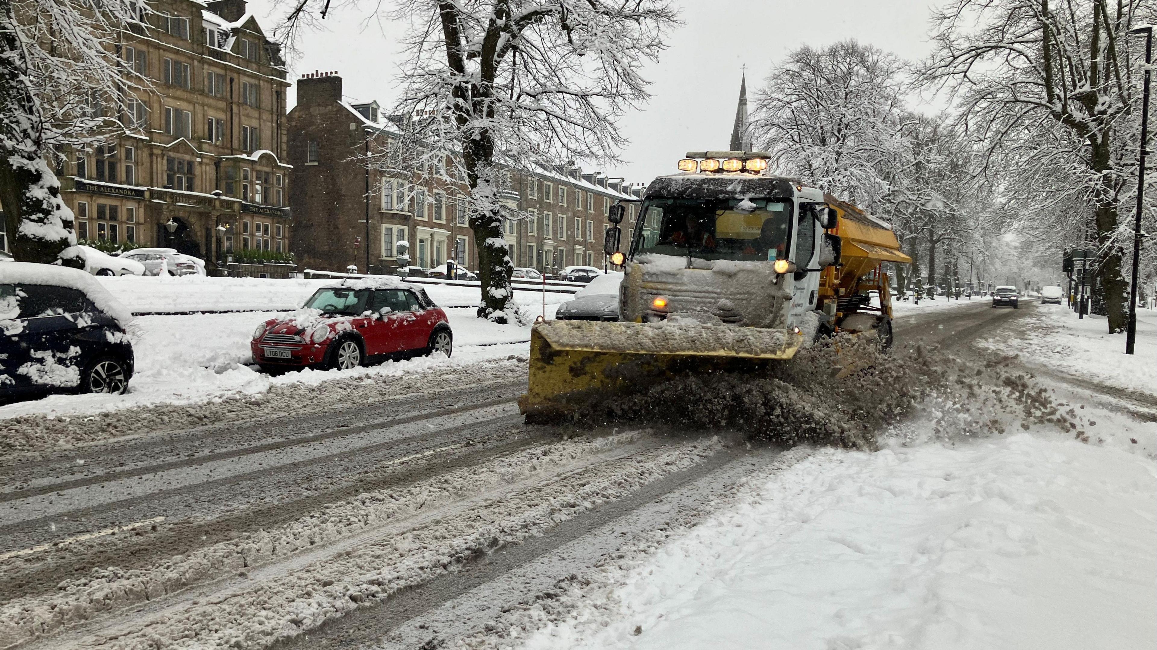 A gritting lorry clears a road in Harrogate with cars parked next to mounted snow on the pavements and hills.