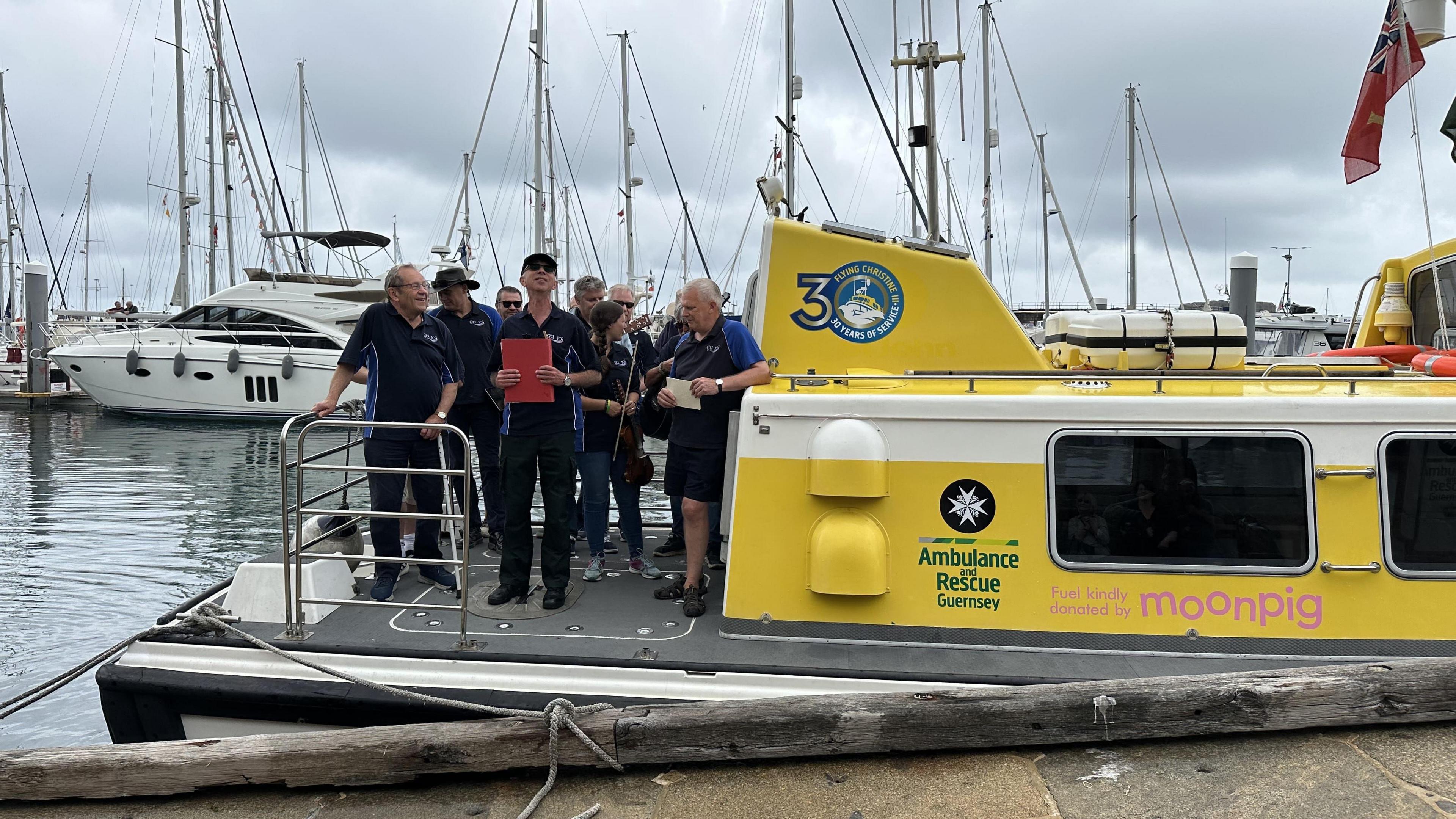 A group of men holding notebooks standing on the back of the Flying Christine III marine ambulance which is docked in a harbour with grey skies behind.