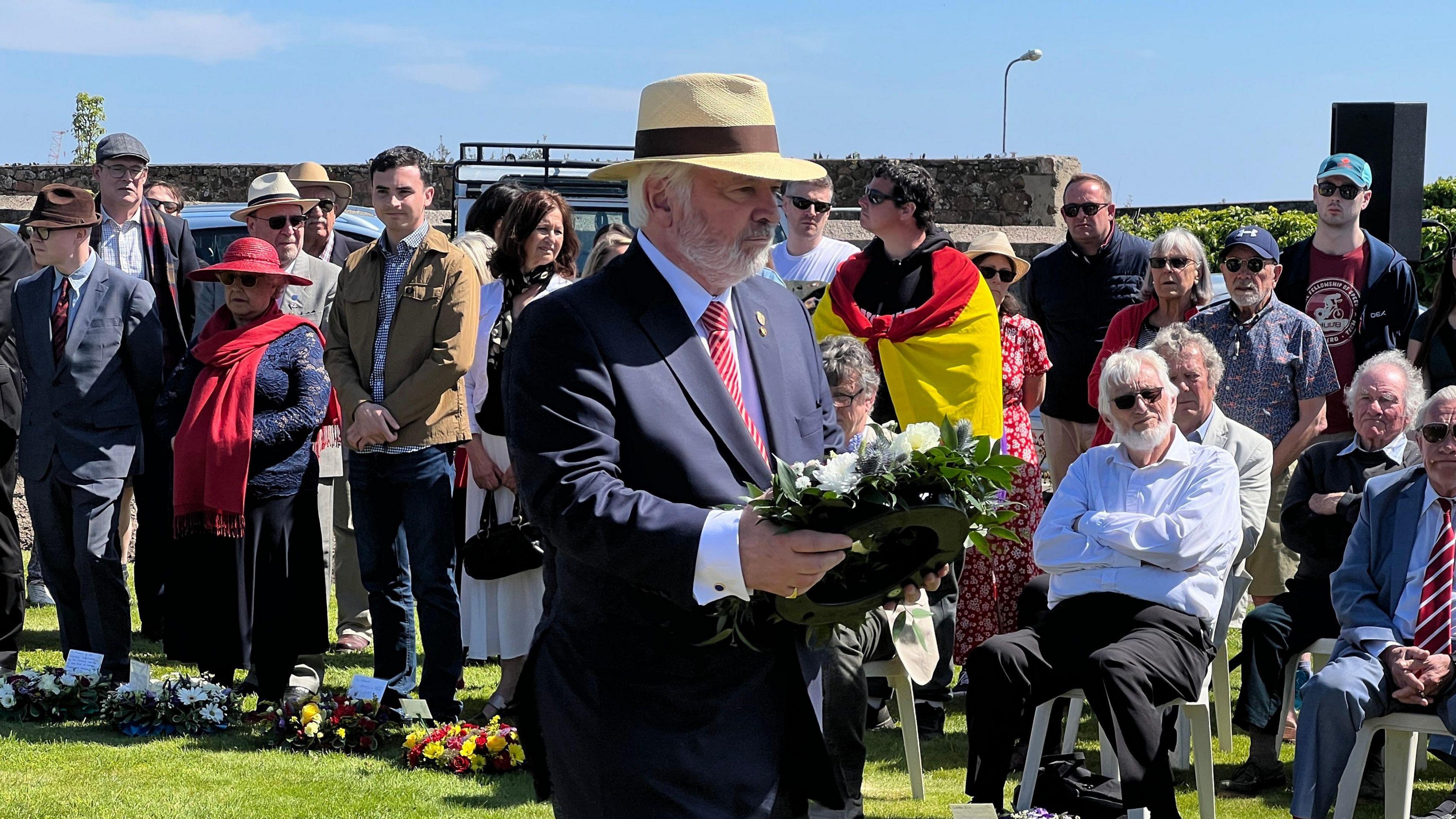 The Bailiff holds a wreath with white flowers as he walks towards to the memorial as a crowd watches behind him