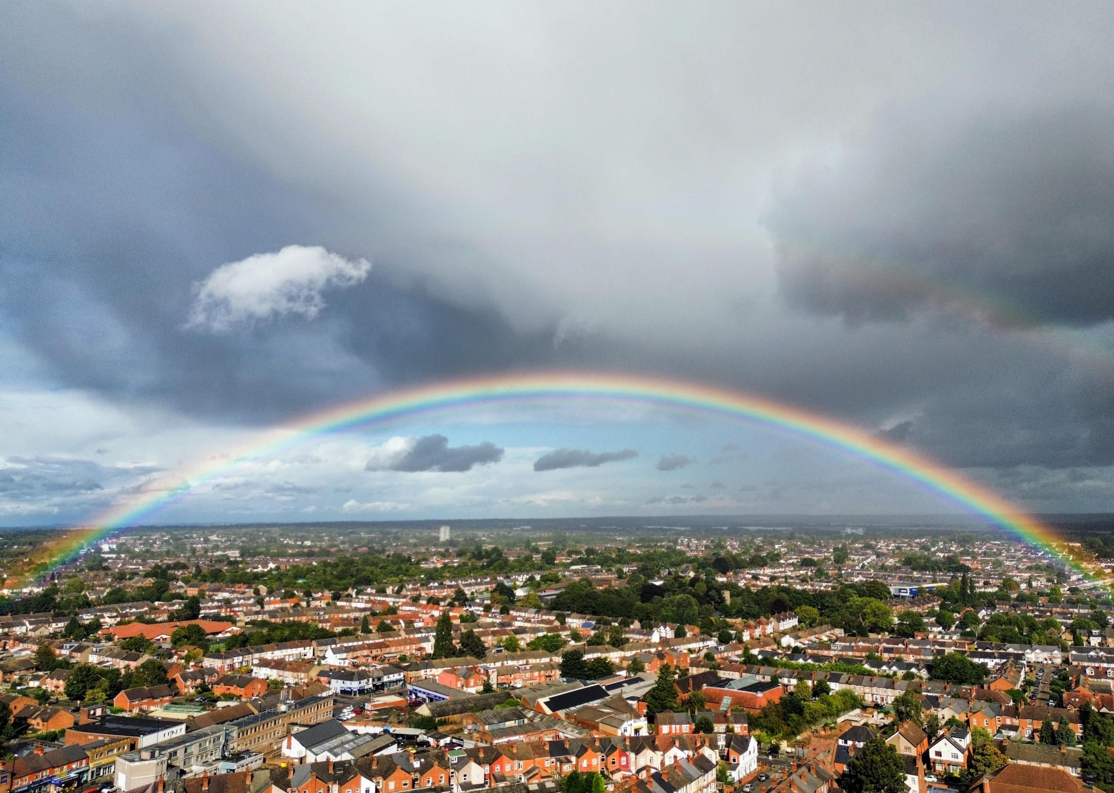 A rainbow spreads across the whole width of a shot taken from high-up over a city suburb. Streets of brick houses with areas of green trees lie underneath, with the horizon several miles away.