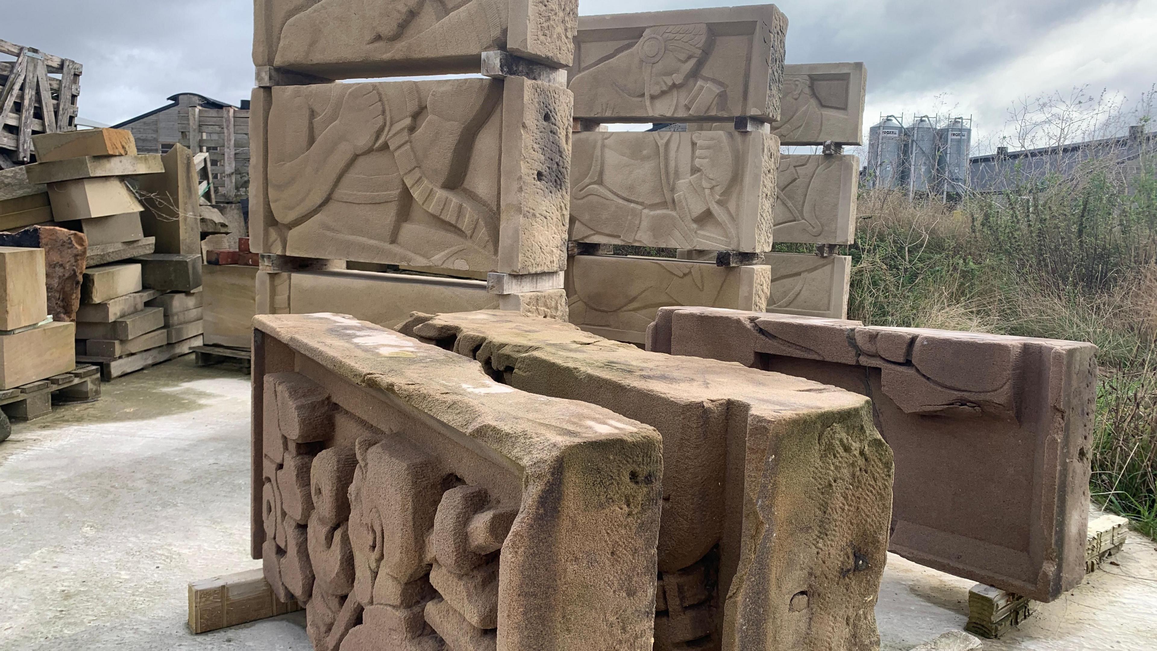 Large pieces of pale stone, with stylised carved figures visible, propped up on pieces of wood within a larger storage area