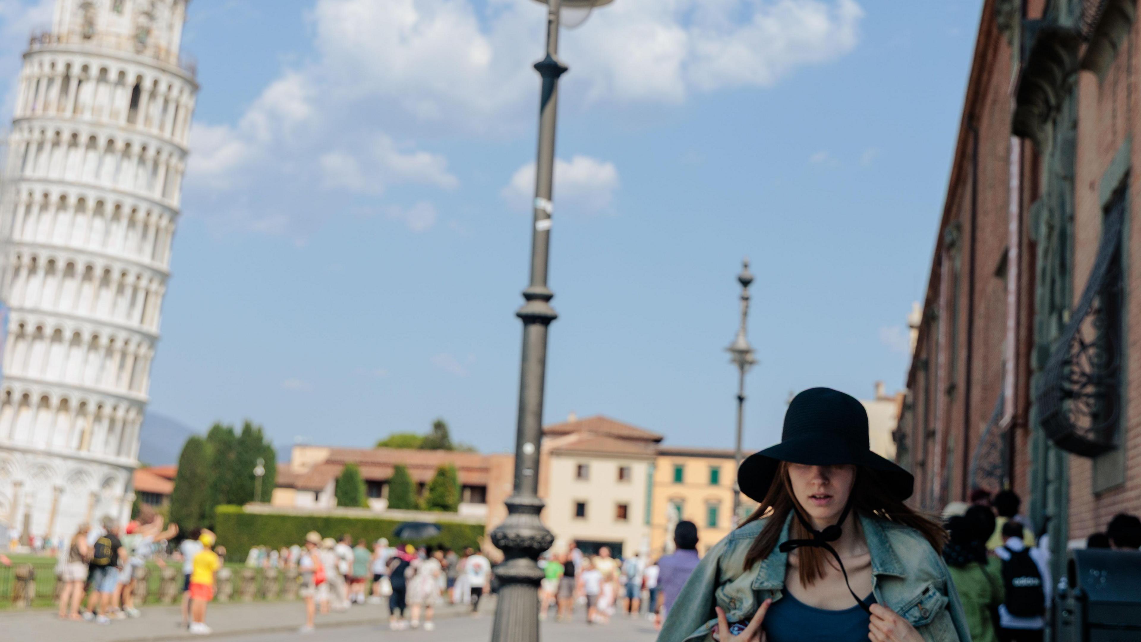 A woman wearing a large sunhat and shielding herself from the sun walks past the leaning tower of Pisa