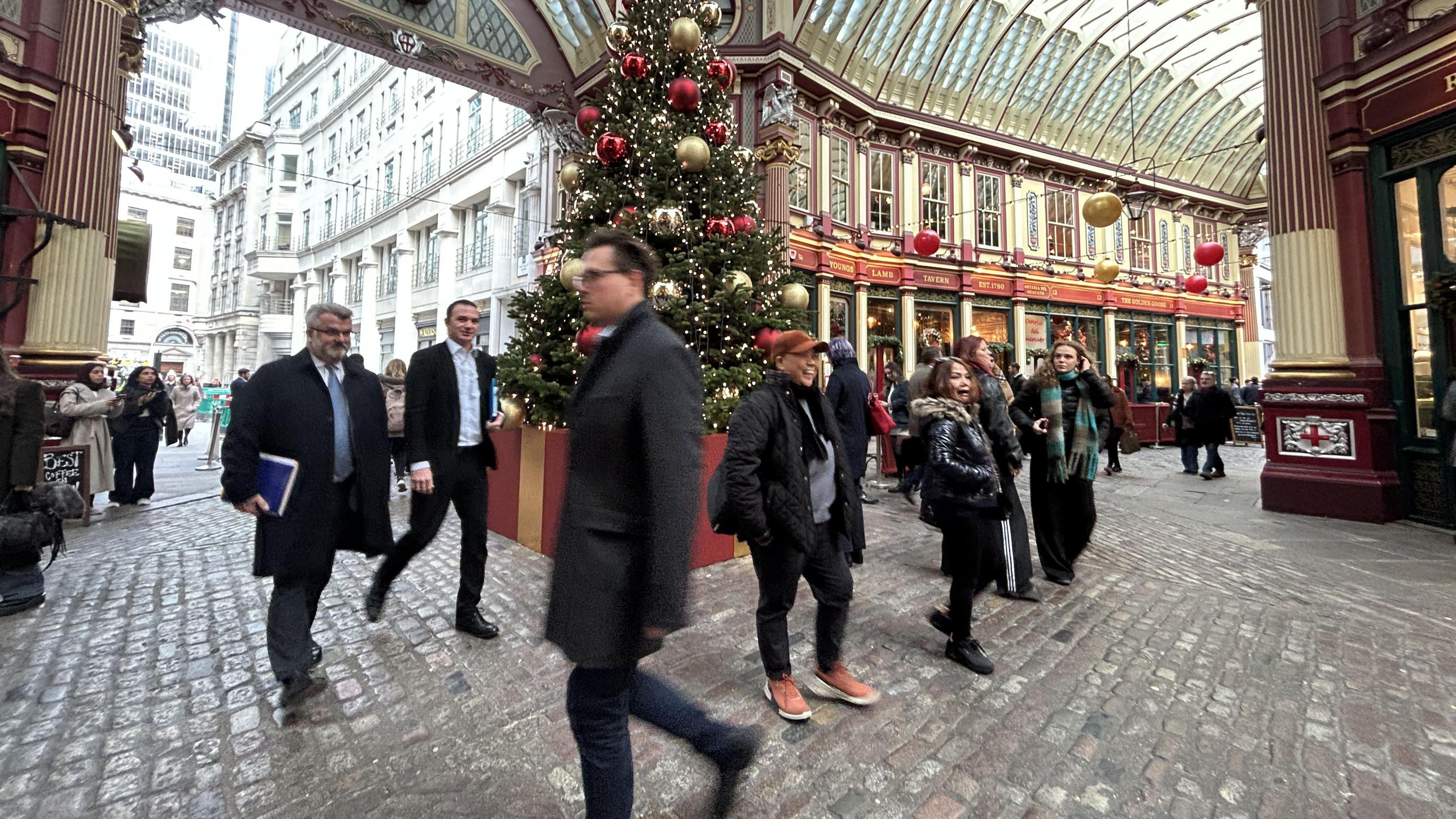 Leadenhall Market with people, including men in suits, walking past a Christmas tree on cobblestone streets. 
