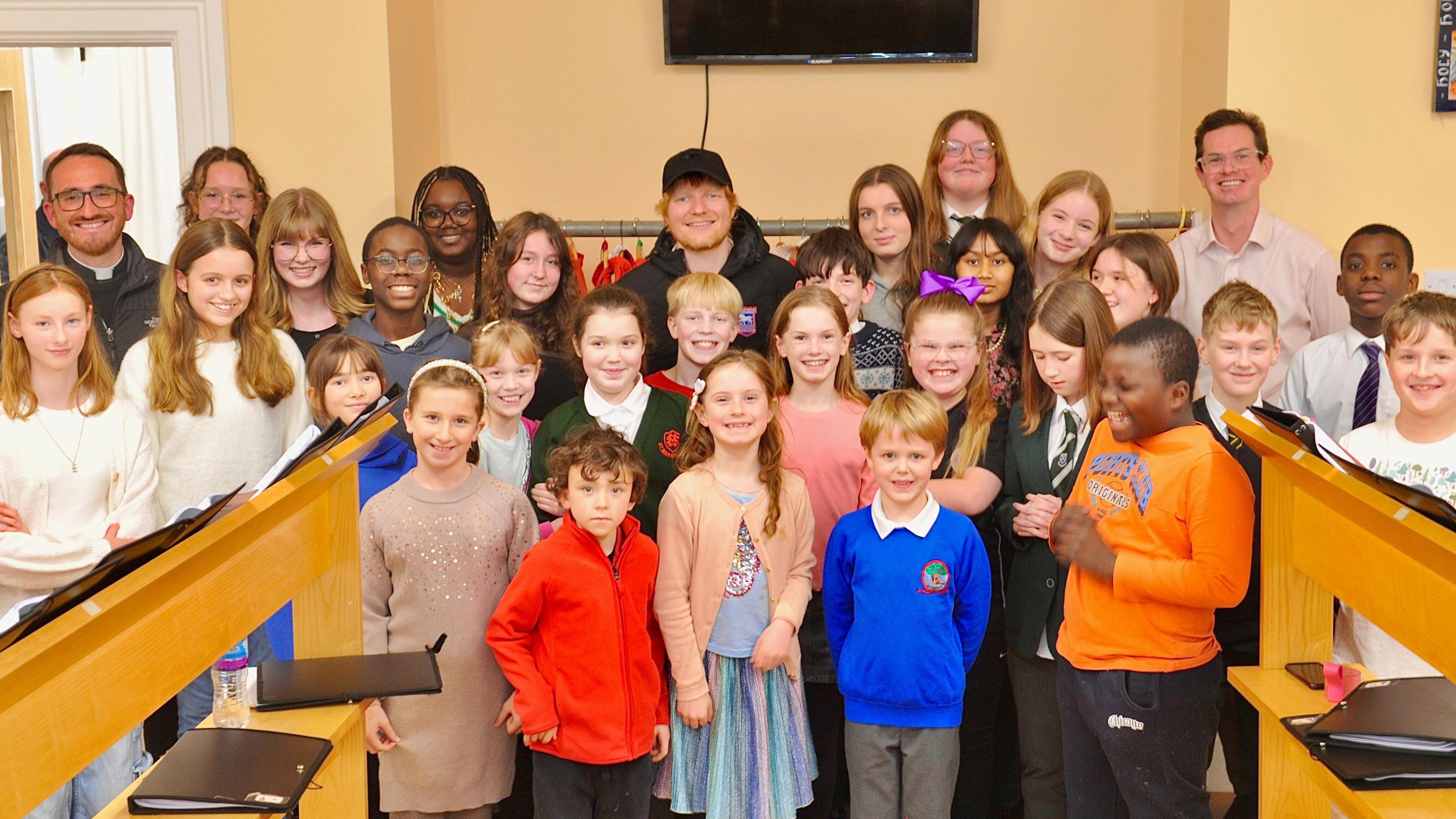 Ed Sheeran standing in a room against a wall, surrounded by about 24 children. He is in the back row wearing a black cap and black jacket with red hair and bead and smiling. There are three other adults, also in the back row