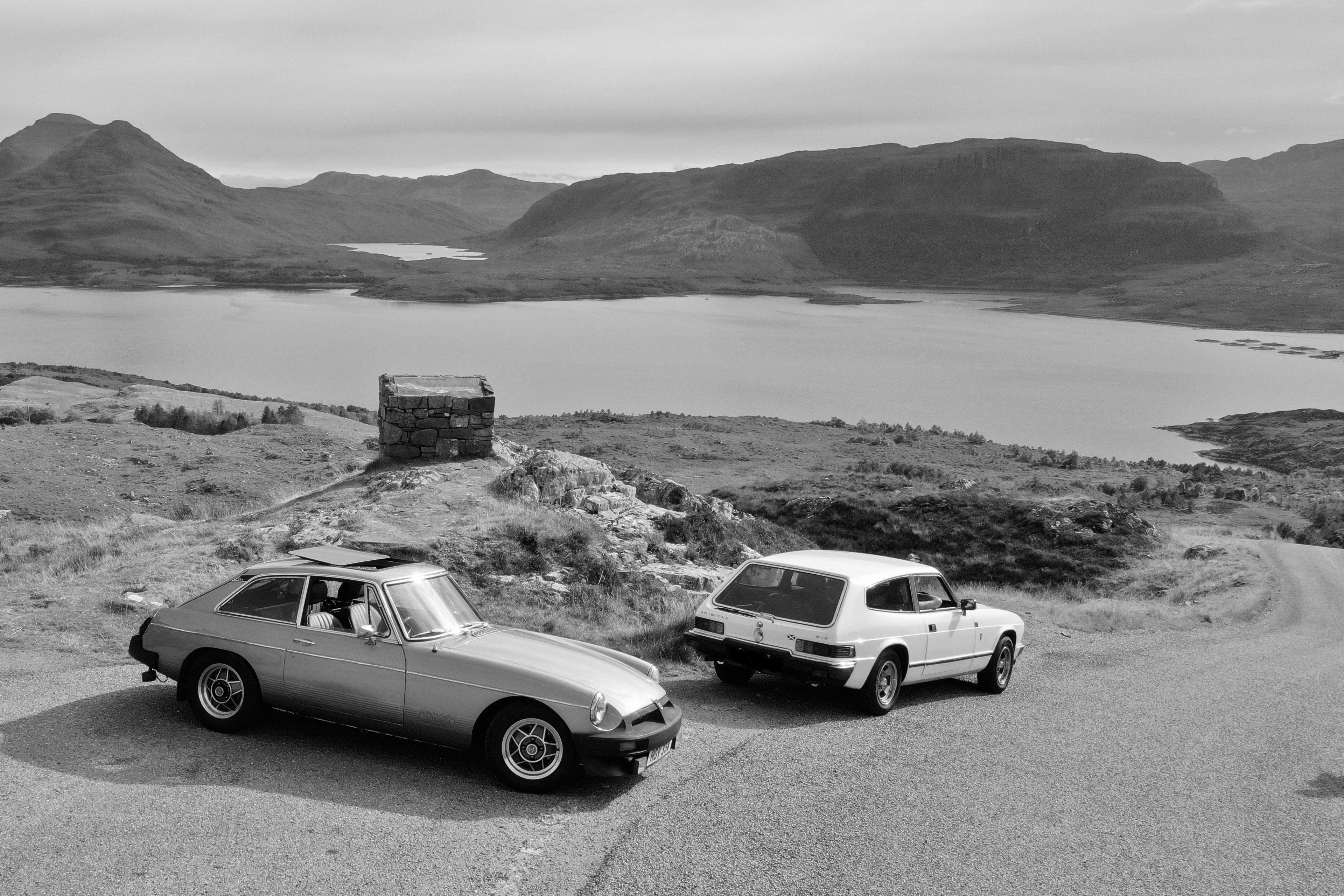 Black and white image of two cars parked on a small hill next to a loch