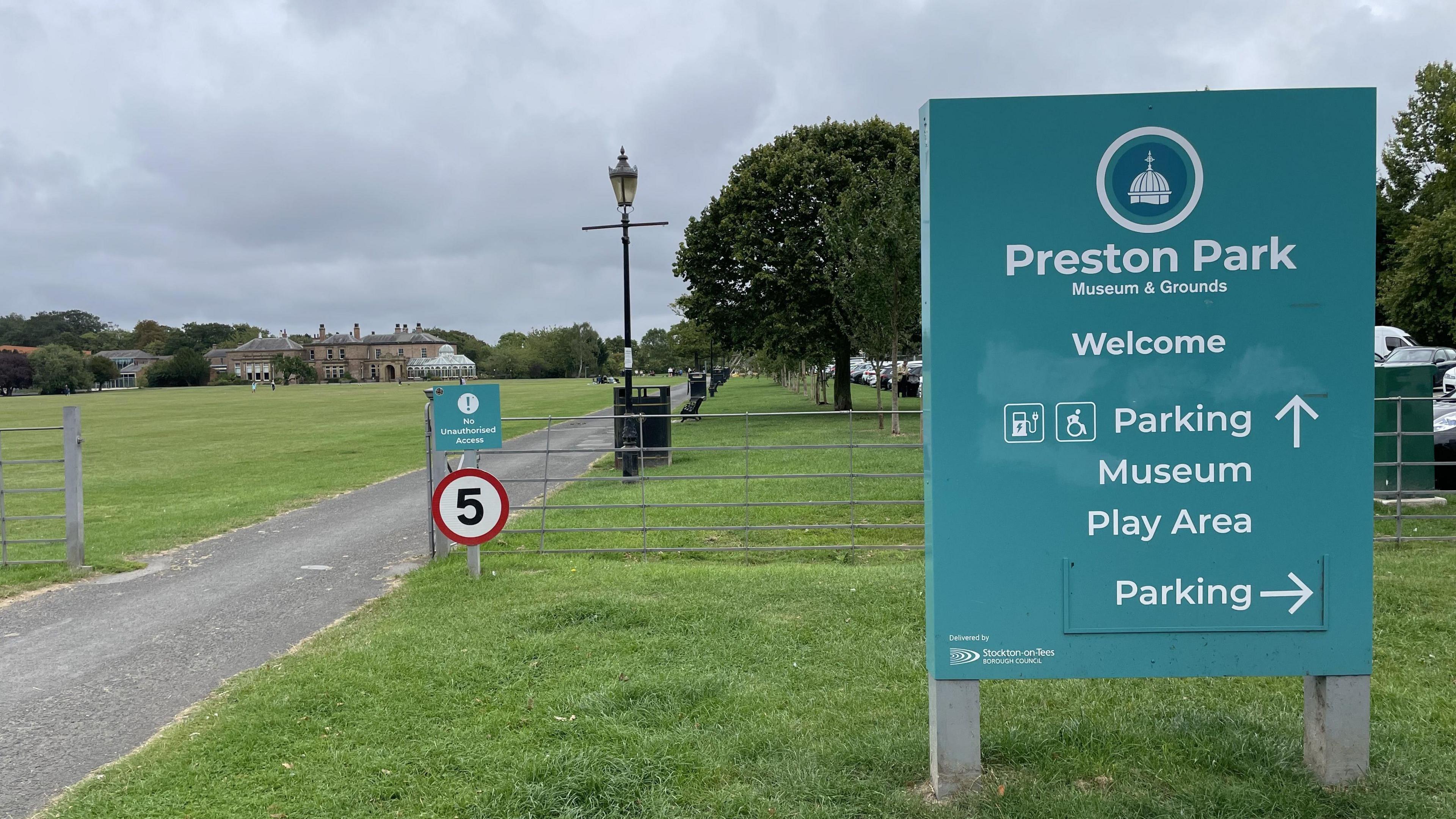 A blue sign with white lettering sits on grass to the right. It reads Preston Park Museum & Grounds and contains directional arrows for parking, the museum and play area. To the left is path that leads up to a country manor style house in the distance. There are trees behind the sign.
