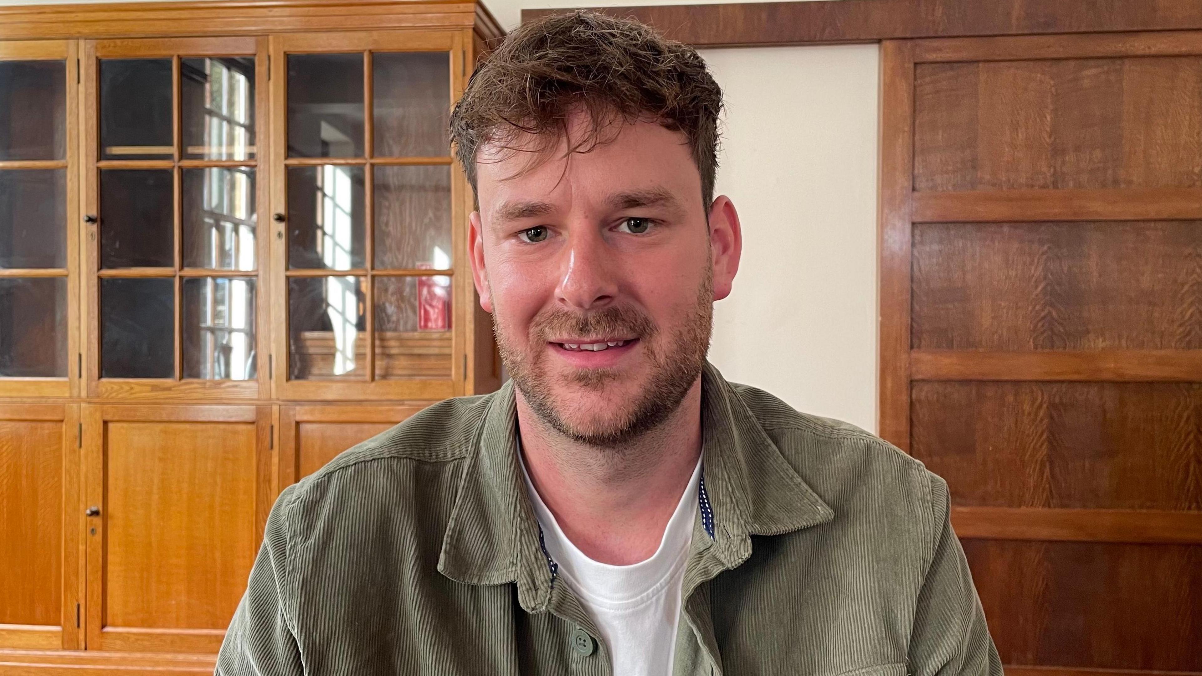 Jack Cooper, looking straight to camera and smiling a little. To the left of the picture is a large empty, glass-fronted bookcase and on the right is a wooden panel. Jack has brown hair and is wearing an unbuttoned sage green corduroy shirt over a white t-shirt.