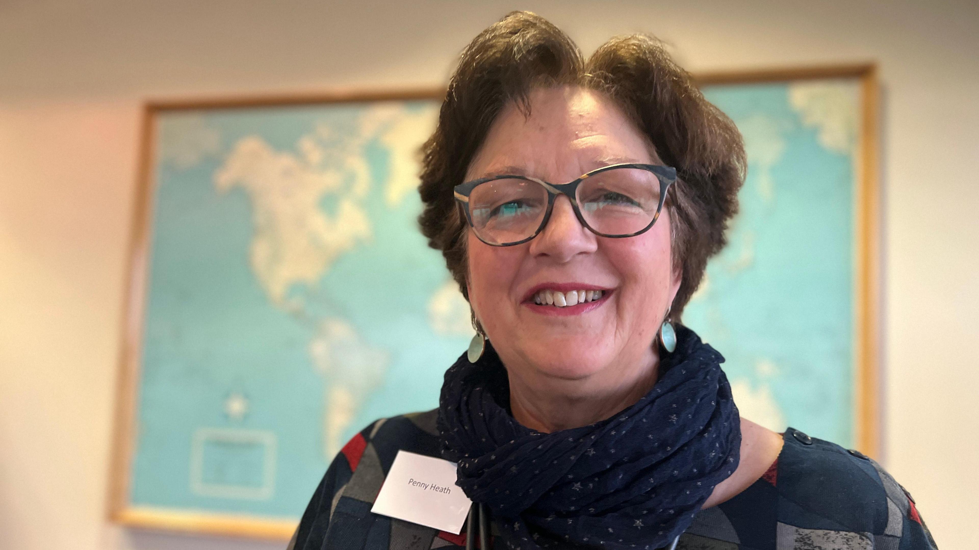 Penny Heath has short brown hair and is smiling at the camera. She is wearing spectacles and a patterned top and blue scarf