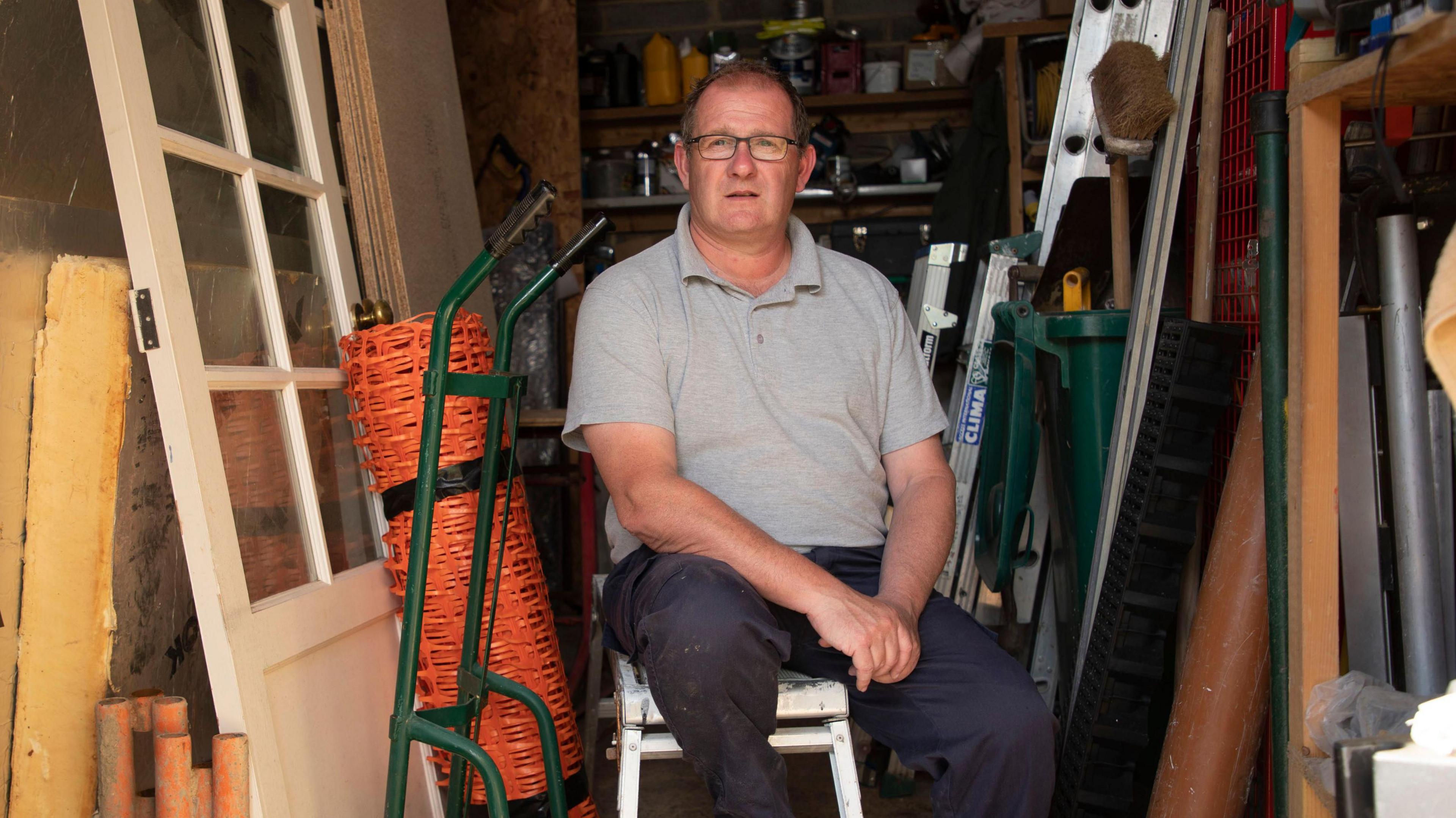 Neil Berriman is wearing glasses and sitting on a metal step ladder in a workshed. He is surrounded by builder's equipment.