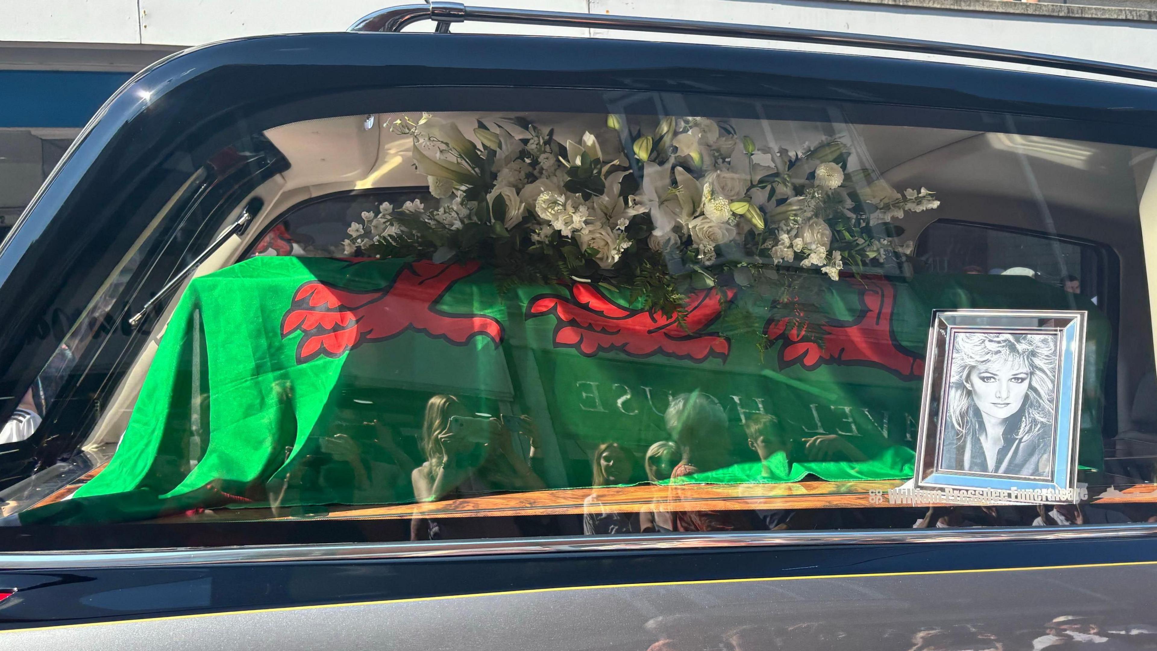 A picture of a coffin in a car. It has a Wales flag over it and has flowers on to. Next to it is a black and white picture of Bonnie Tyler