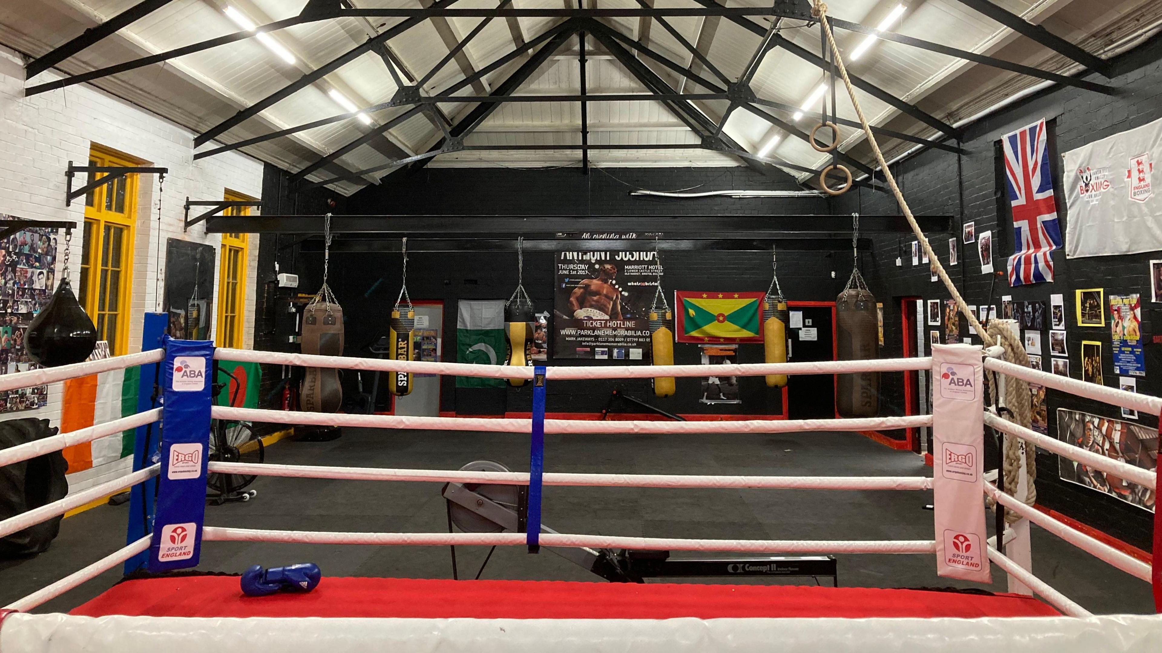 A boxing ring in the foreground, and various punch bags around the wall of the sports club