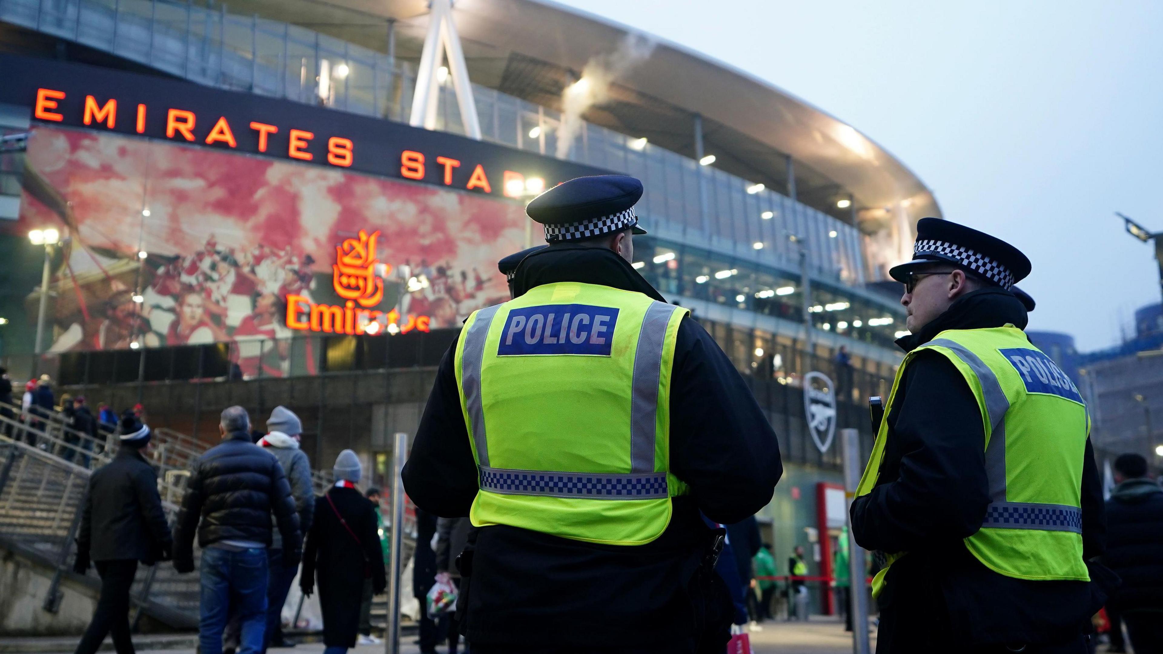 Two police officers with their back to the camera outside Arsenal's stadium watching fans making their way into the ground. 