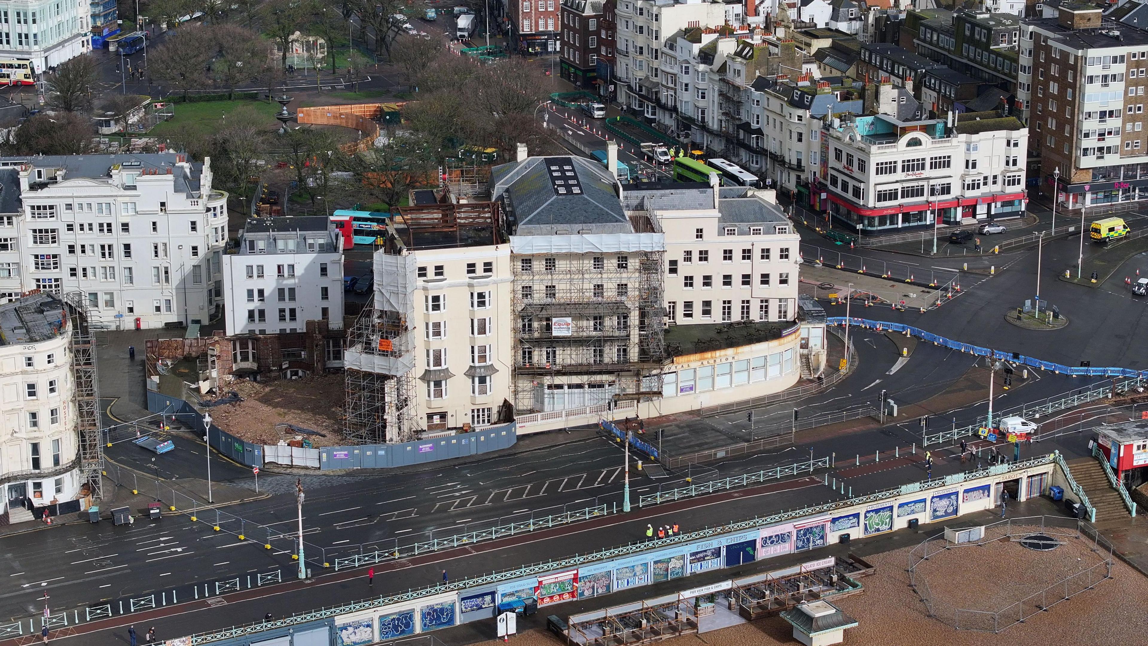 An aerial shot showing a section of blue barriers across the road which leads to the hotel. The Royal Albion can be seen covered in scaffolding in the centre of the image. The beach is just visible in the foreground.