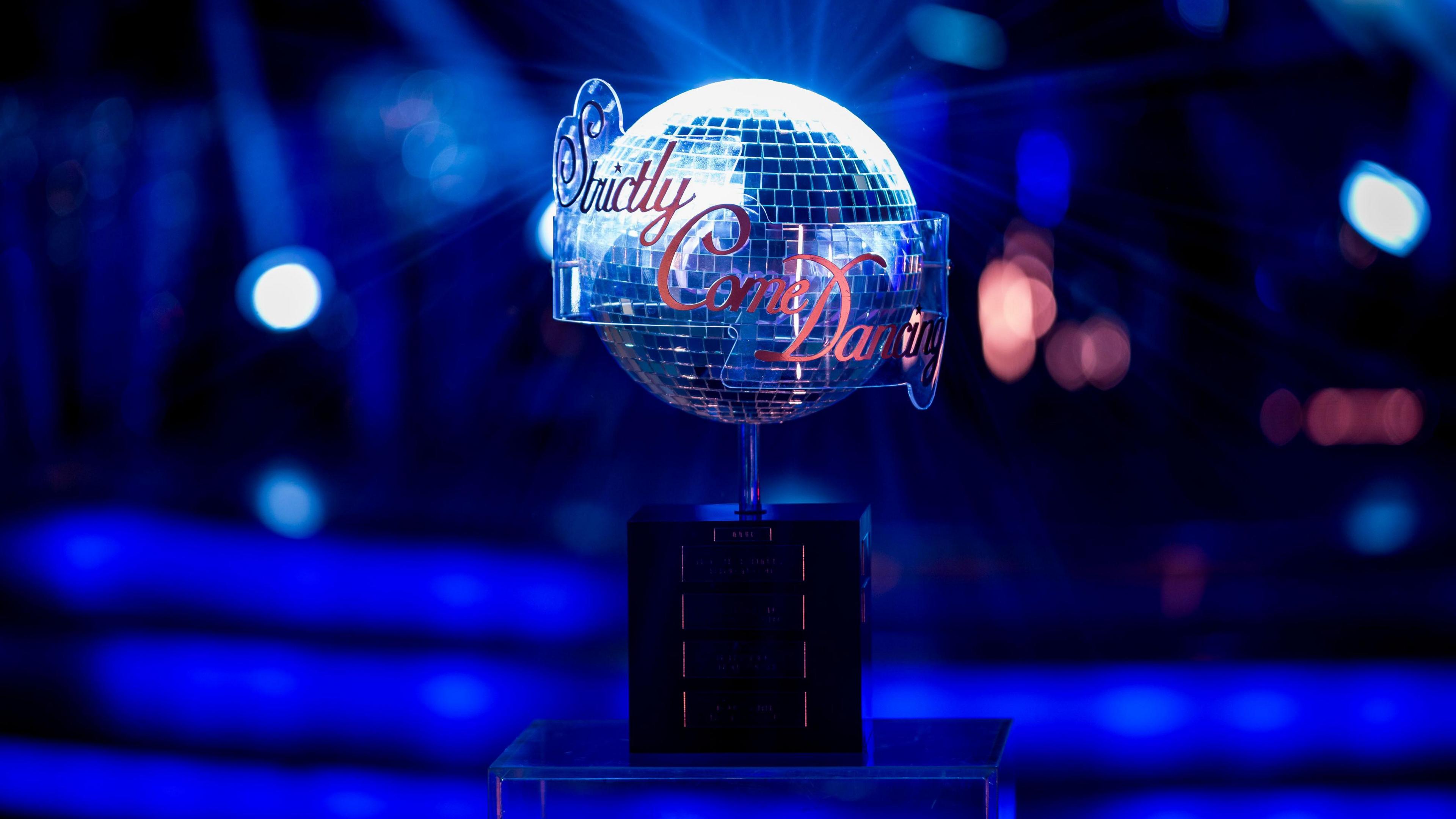 The Strictly Come Dancing sits on a black plinth against a brightly lit blue studio background which is blurred