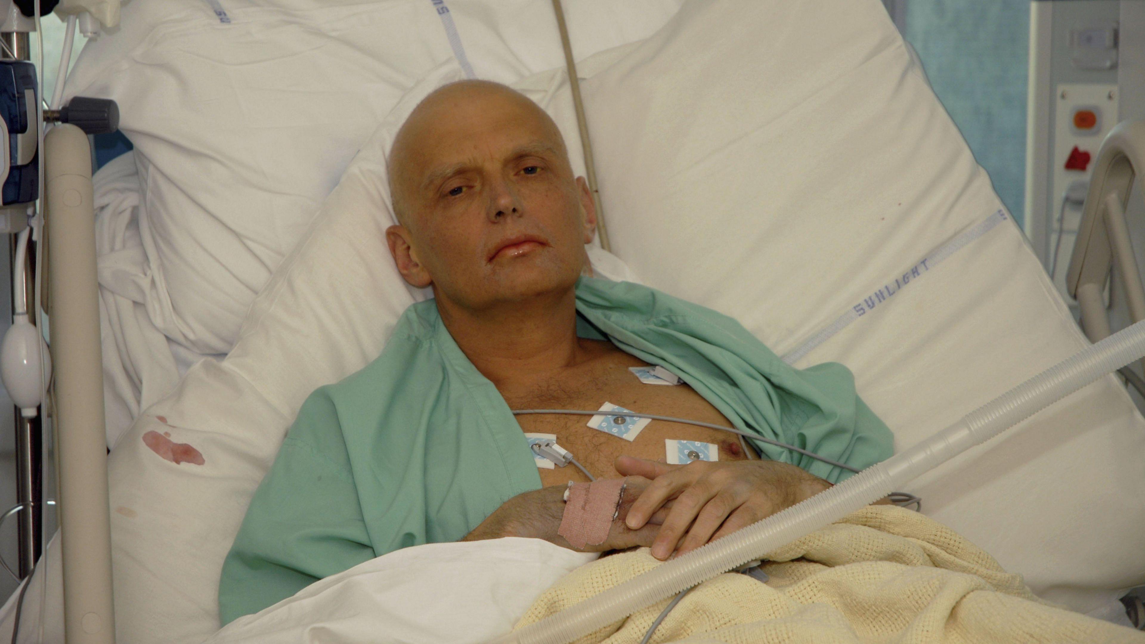 Alexander Litvinenko lying in a hospital bed on an intensive care unit. Lots of wires are linked up to his chest. He has a bandage on one hand. He is looking into the camera with a blank expression on his face. 