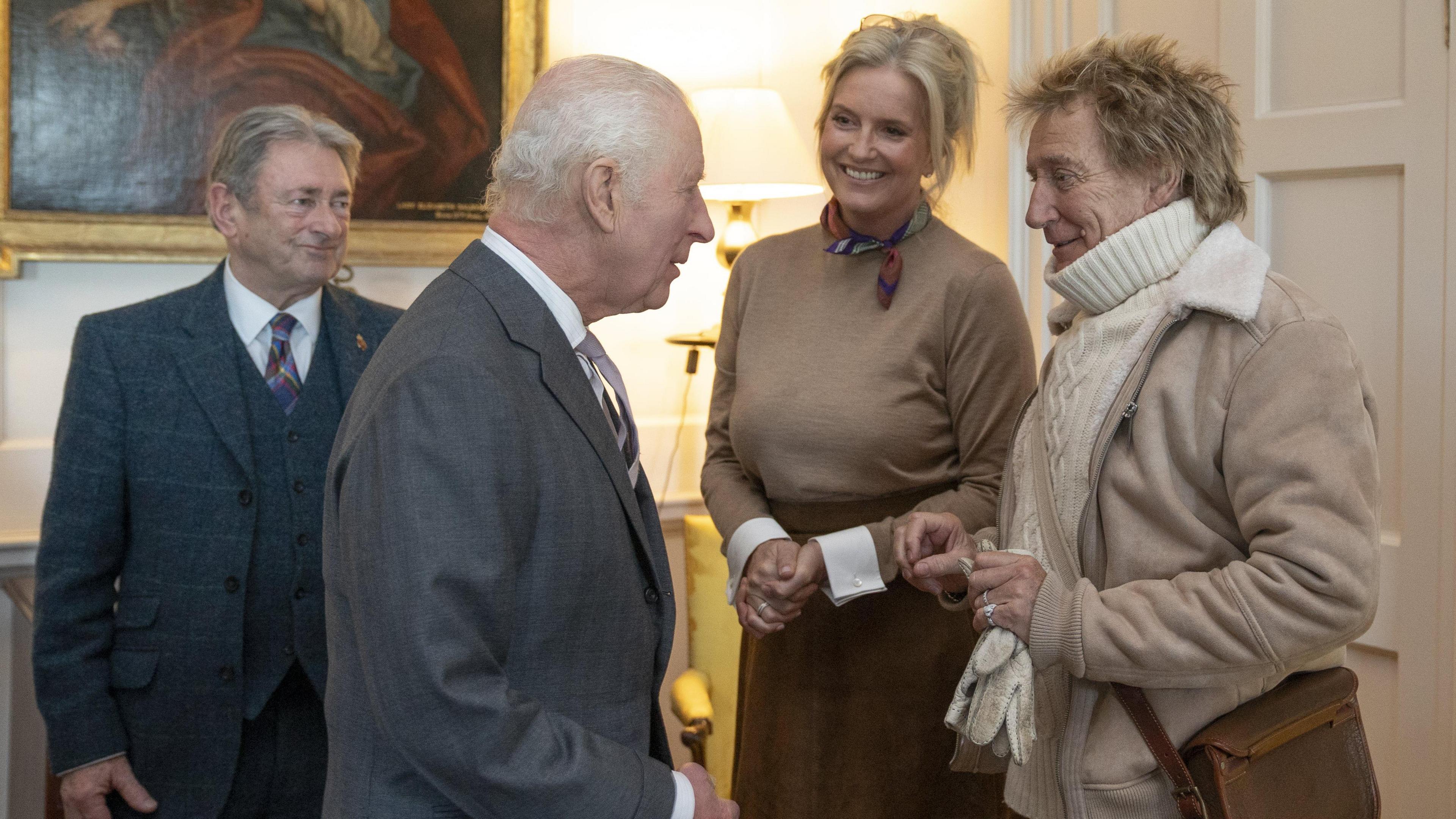 King Charles, wearing a grey suit with a white shirt and light tie, talks to Sir Rod Stewart, wearing a white turtleneck jumper under a beige jacket and with a beige strap covering his torso, and wife Penny Lancaster. Alan Titchmarsh, wearing a dark suit with a dark multi-coloured tie, looks on.