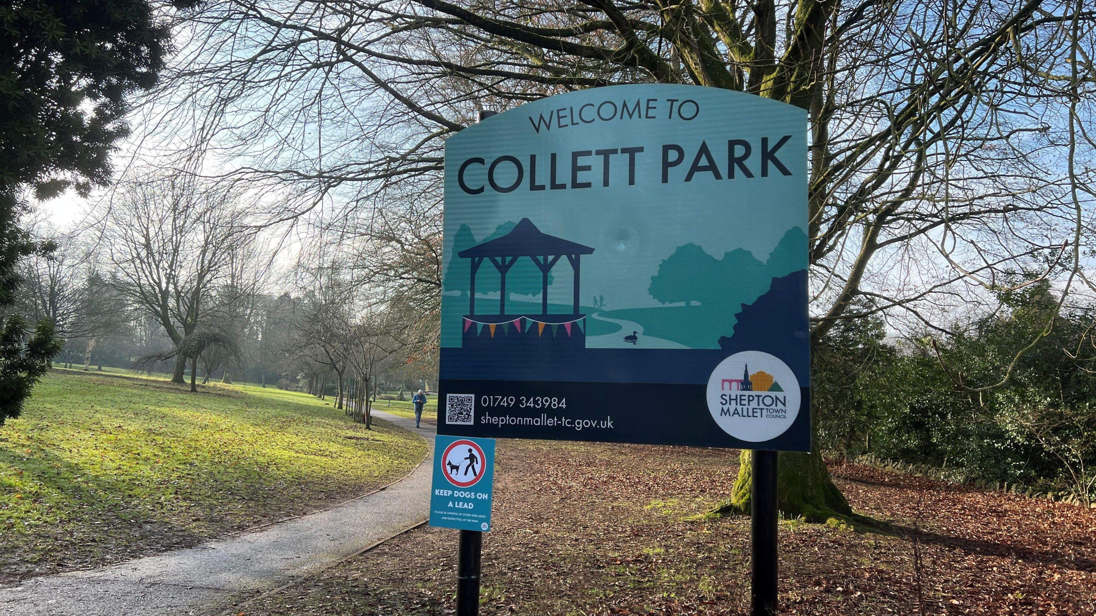 Picture of park with green spaces. There's a blue sign saying "Welcome to Collett Park" in big writing. At the bottom of the sign it says "Keep Dogs on a lead" with a big red circle with a dog walker.