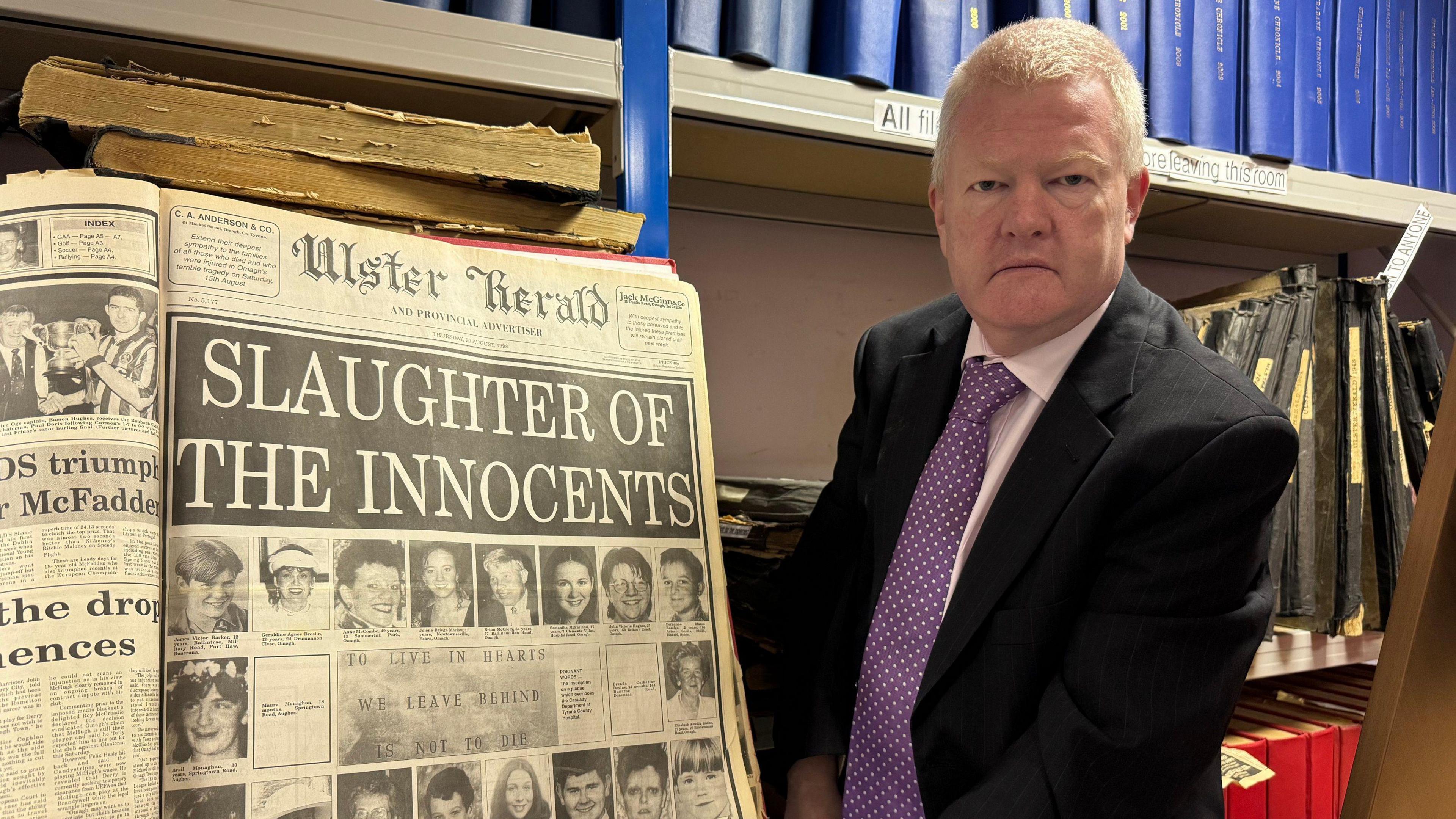 Alan Rodgers. He has short white hair, wearing a black blazer, pink shirt and purple polka dot tie. He is holding an old Ulster Herald newspaper, the page says 'Slaughter of the Innocents' with pictures of the victims.