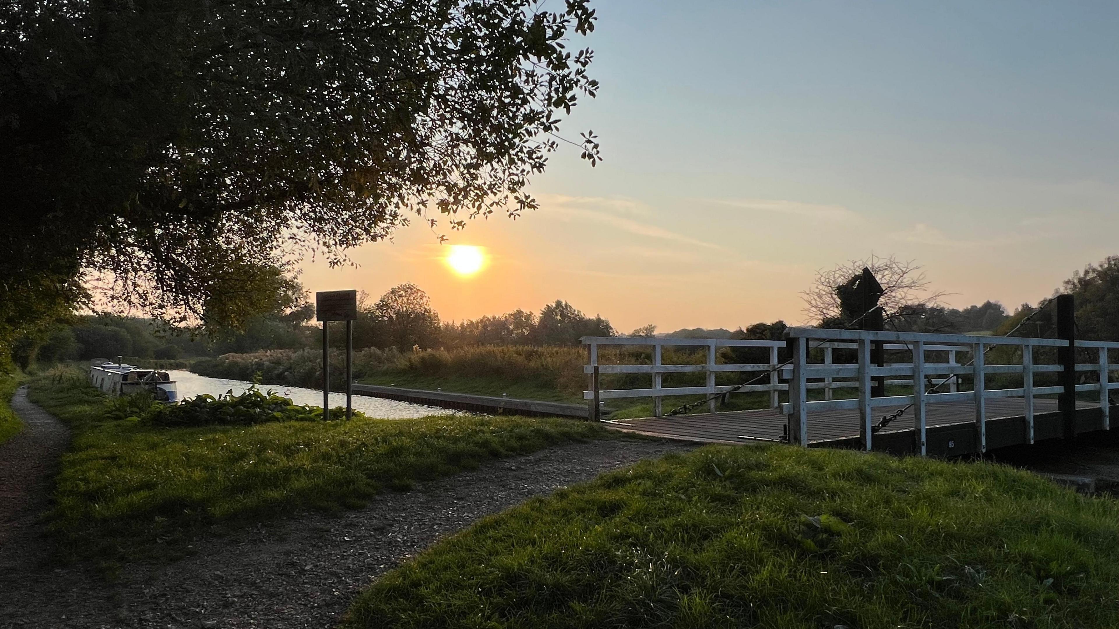 The sun sets on a clear evening. A fence-lined bridge can be seen on the right-hand side of the image and a grassy area with overhanging trees on the other side. 