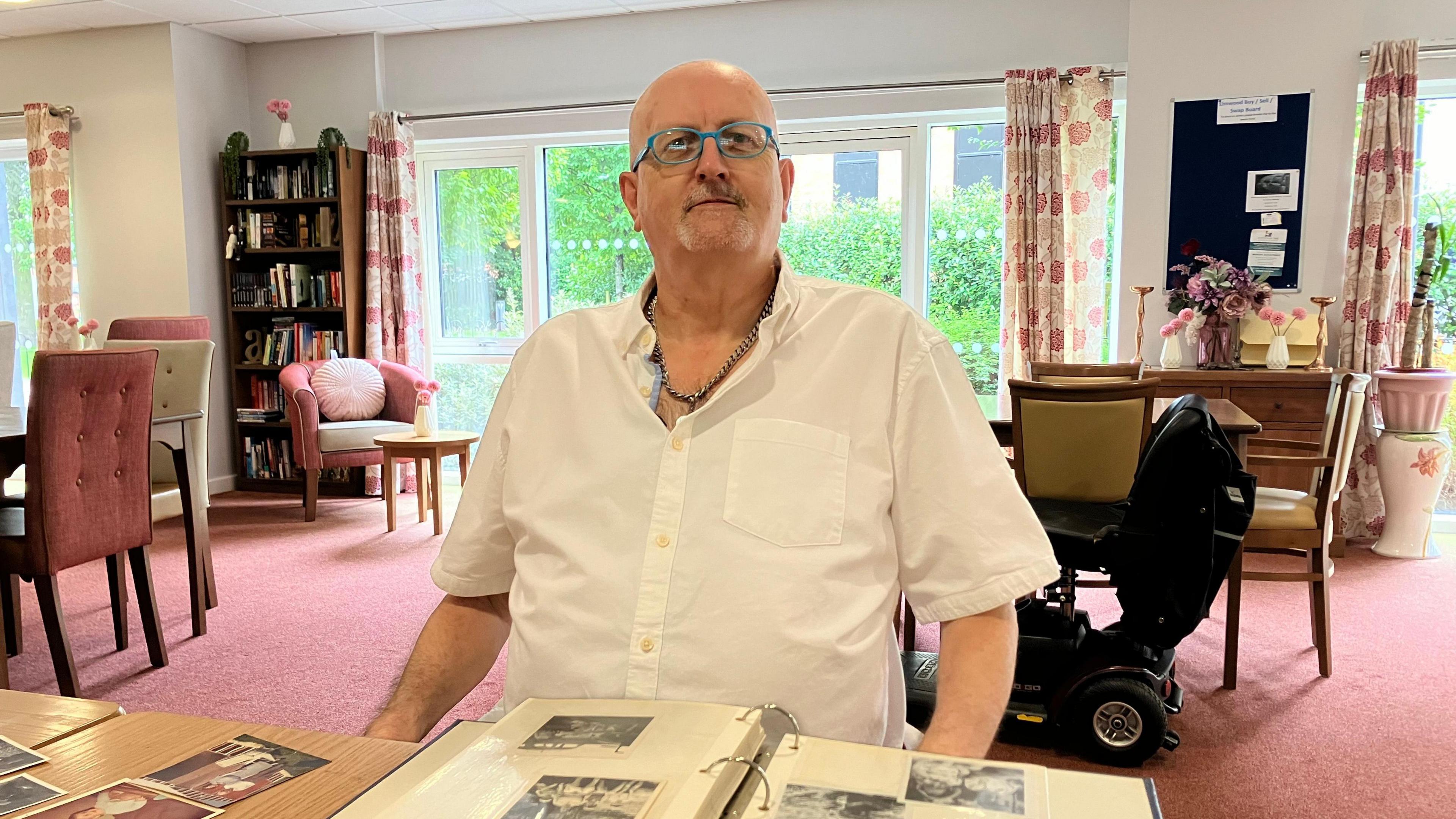 Bald man wearing glasses and a white shirt is sat in front of a photo album, inside a lounge with a pink carpet and various pieces of furniture, as well as a mobility scooter 