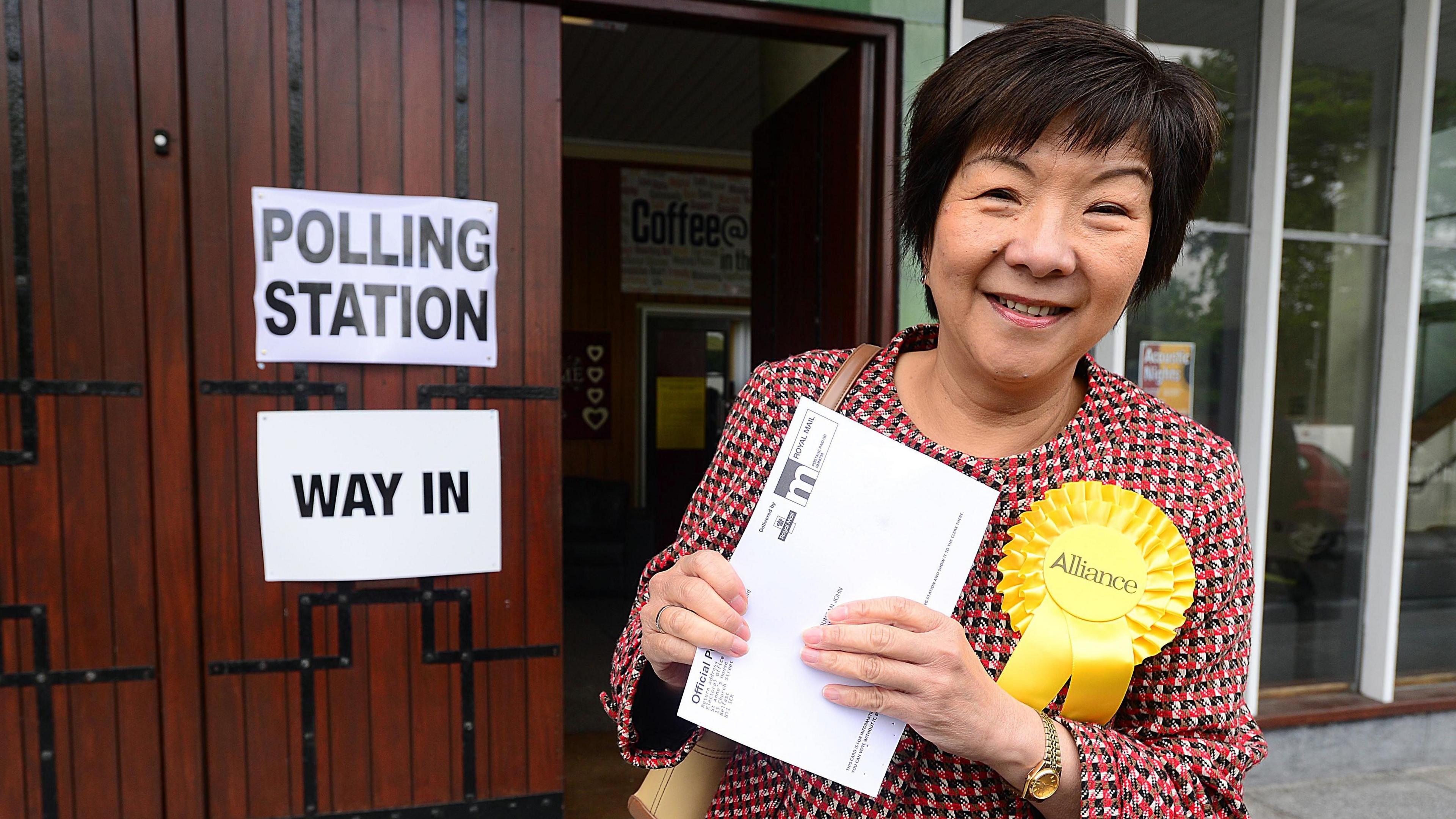 Archive photo of Anna Lo pictured outside a polling station.  She is wearing a multicoloured jacket and a yellow Alliance rosette.  She is smiling and holding a polling card. 