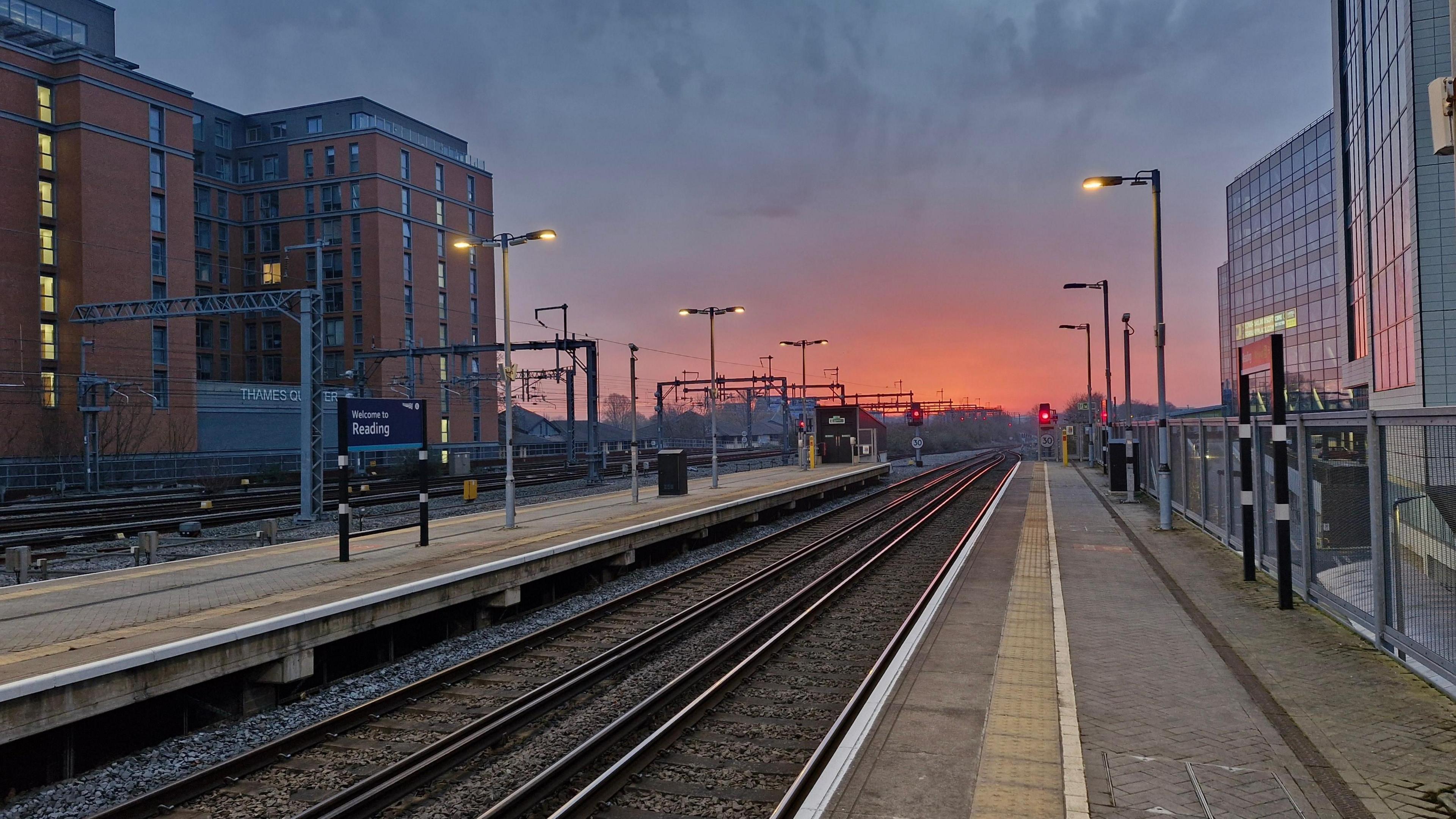 Sunrise as seen from Reading train station. Tracks run off into the distance where an orange glow can be seen as the sun rises. The platforms are deserted in this early-morning shot.