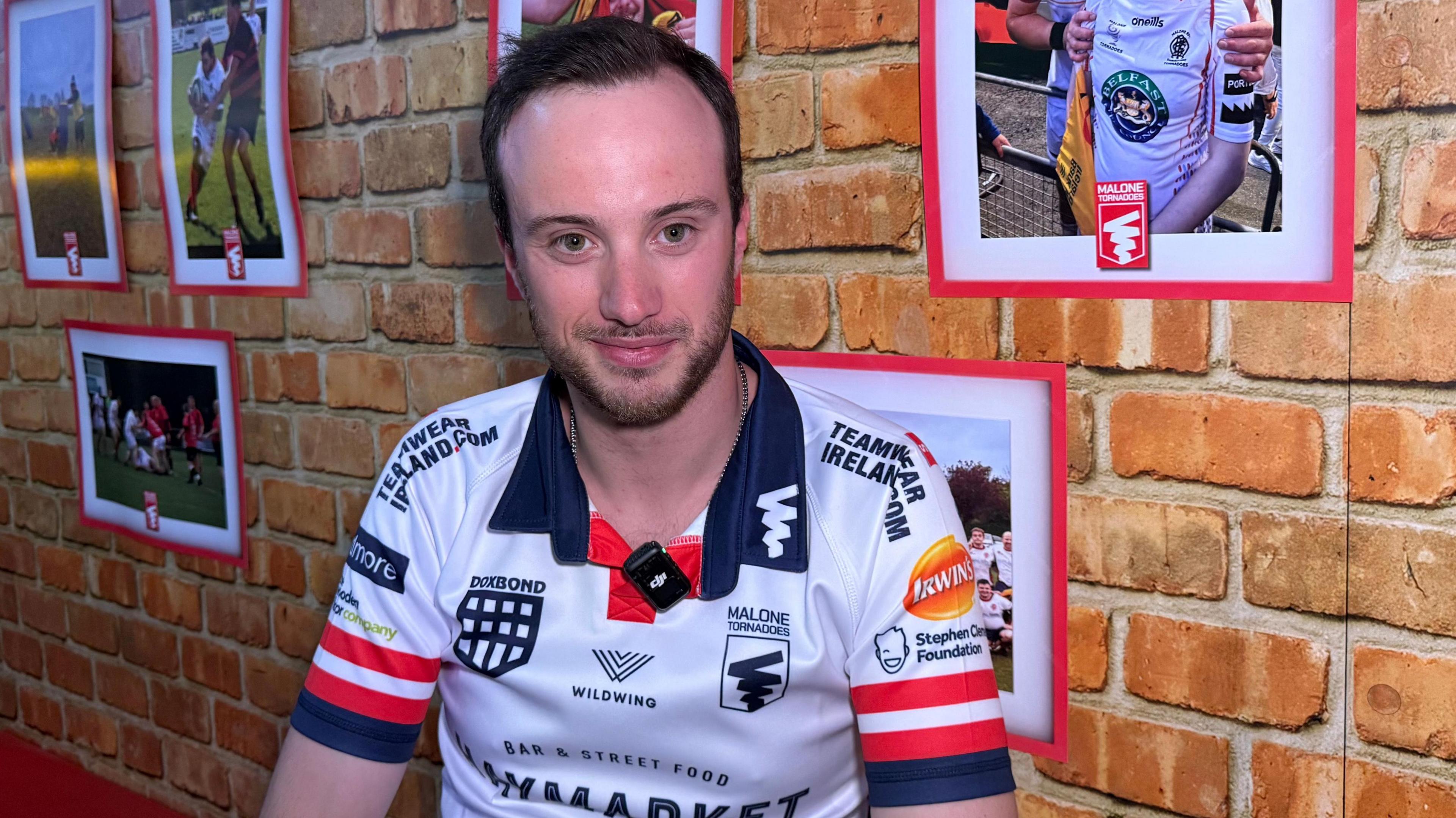 A brown haired man with blue eyes sitting down in front of a brick wall with rugby photos on it. He is smiling and has a beard.