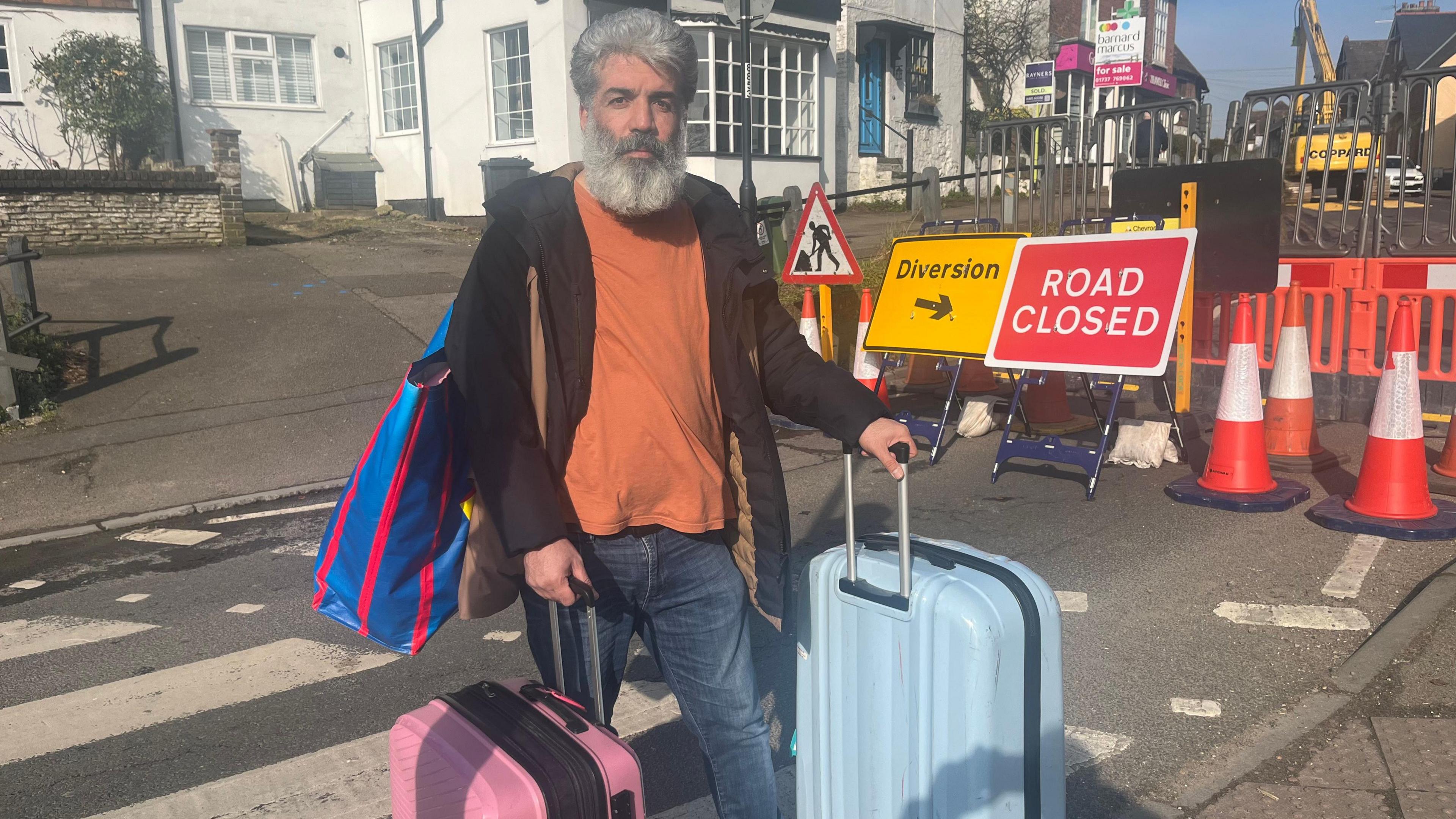 A man with a large grey beard wheels two suitcases and carries a bag over his shoulder, as he leaves his home in Godstone. He wears a brown hoodie, orange t-shirt and blue jeans and has grey hair.