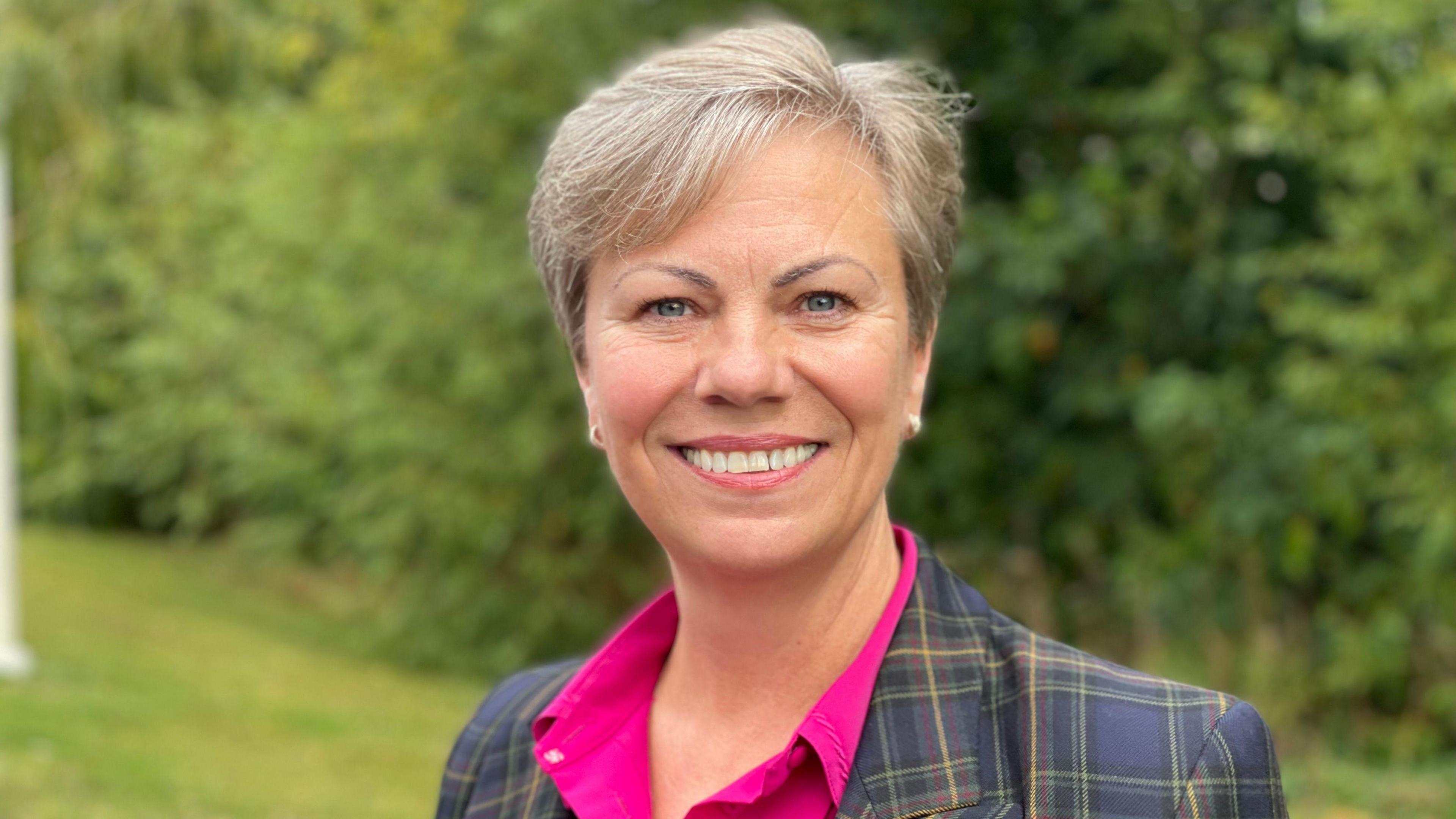 Dawn has short grey blonde hair and is wearing a fuchsia pink shirt with a navy and green tweed patterned suit jacket on top