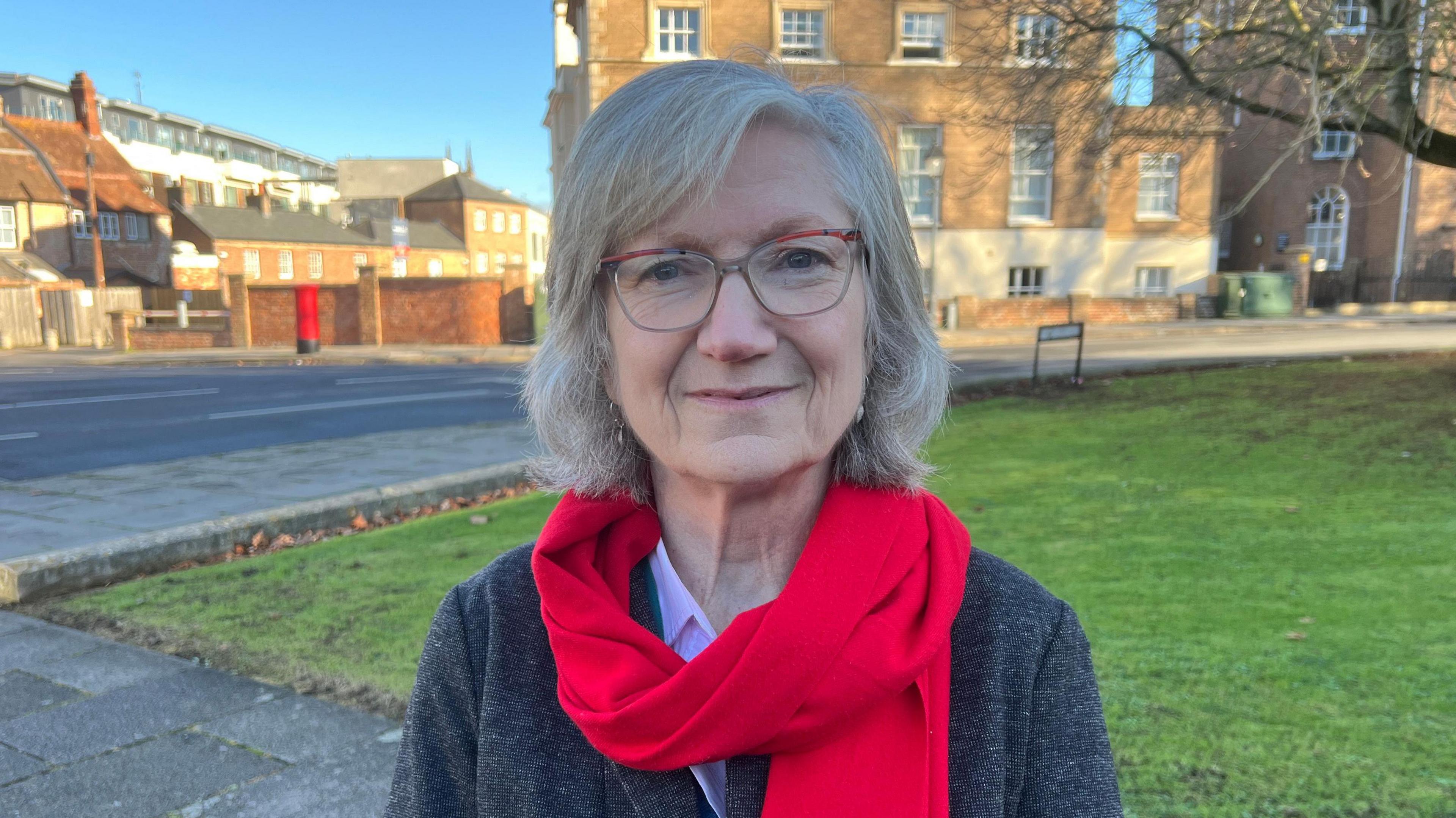 Women with grey hair and glasses standing outside near a patch of grass and some buildings. She is wearing a red scarf and wearing a dark coat.
