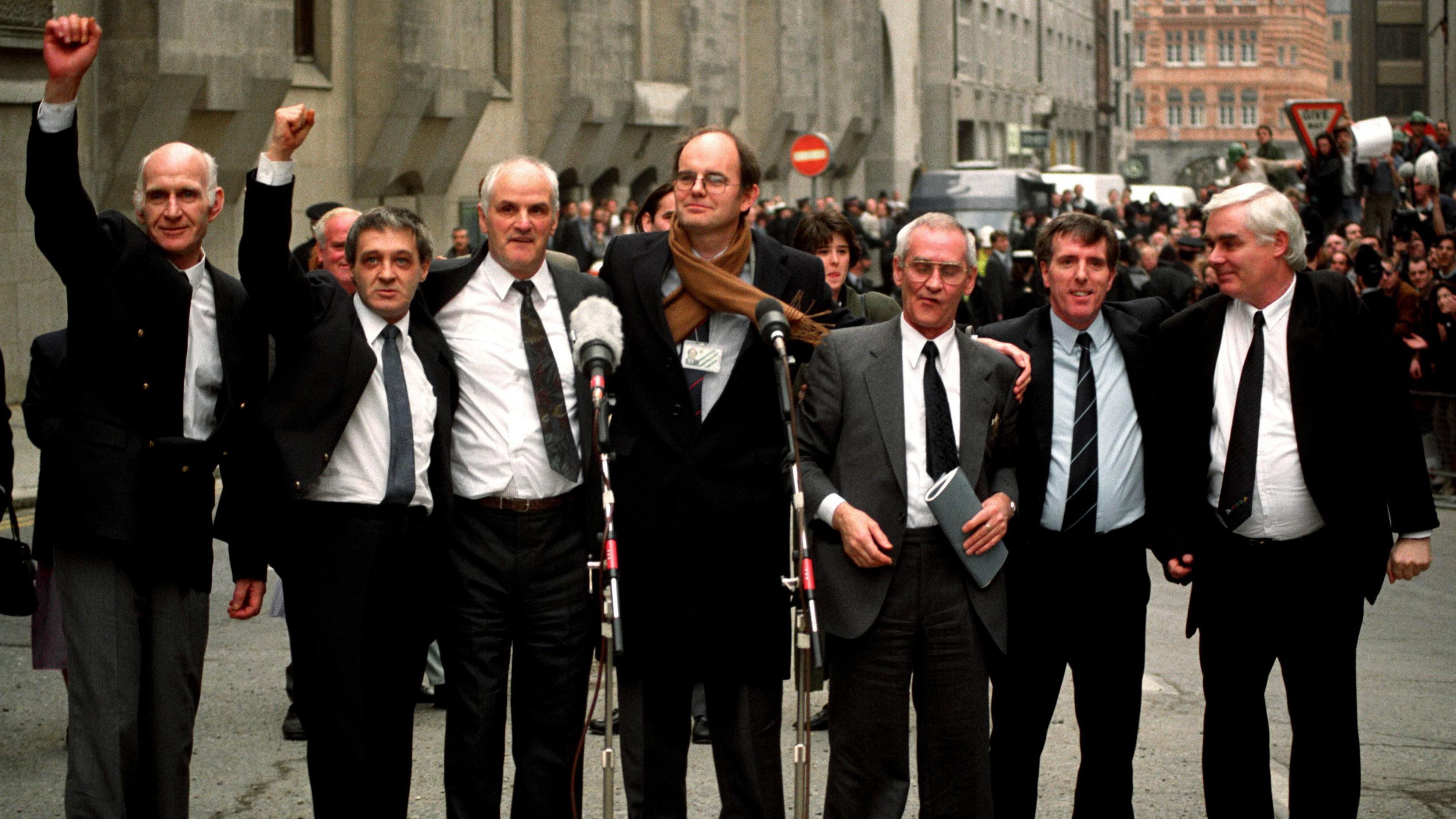 Left to right is John Walker, Paddy Hill and Hugh Callaghan; the then MP Chris Mullin; Richard McIlkenny, Gerry Hunter and William Powe. All wear suits. 