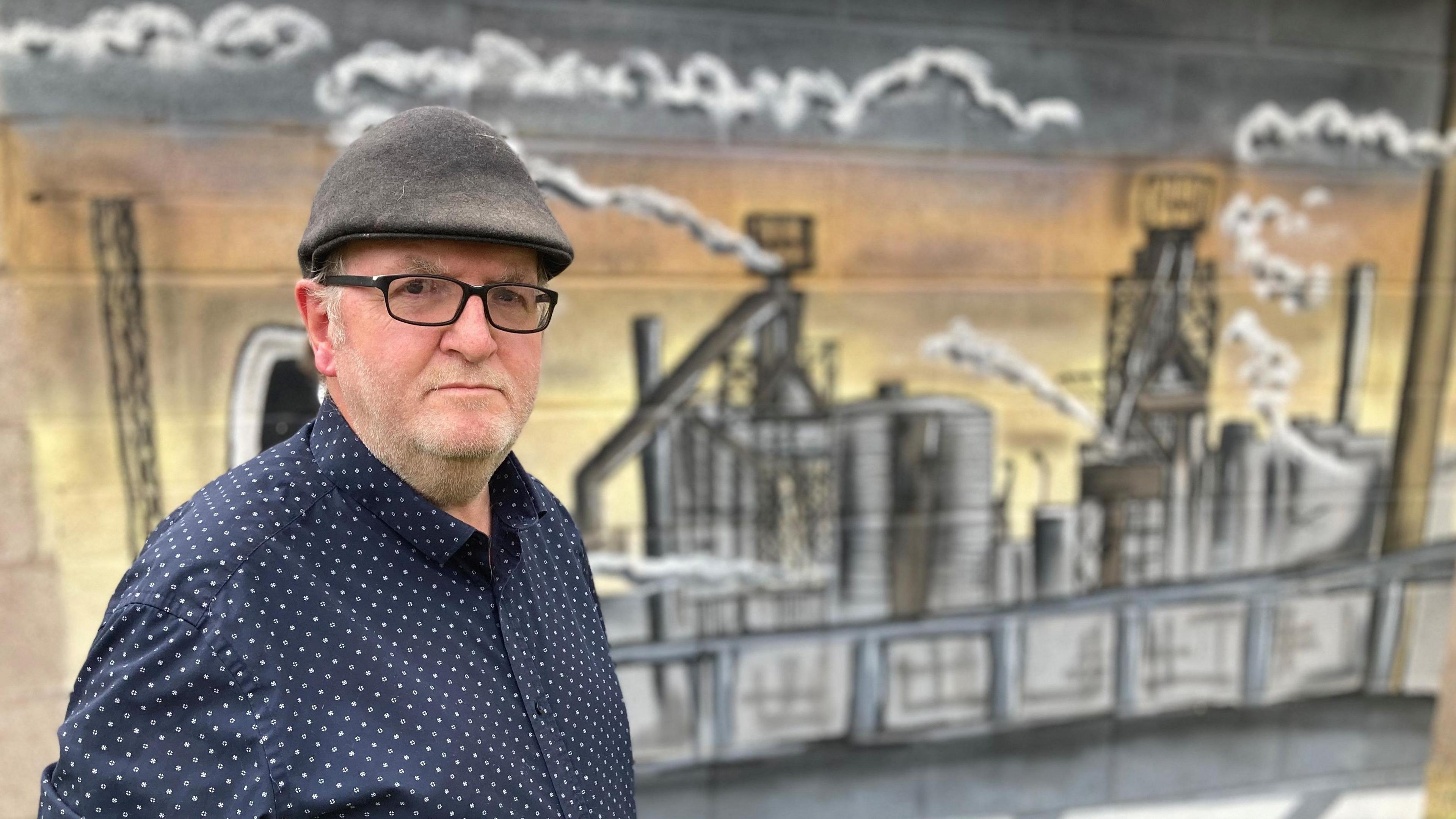 Gary Owen looking at the camera. He is wearing a blue spotted shirt and a flat cap, he has glasses on. He is stood in front of a piece of artwork depicting the steelworks.