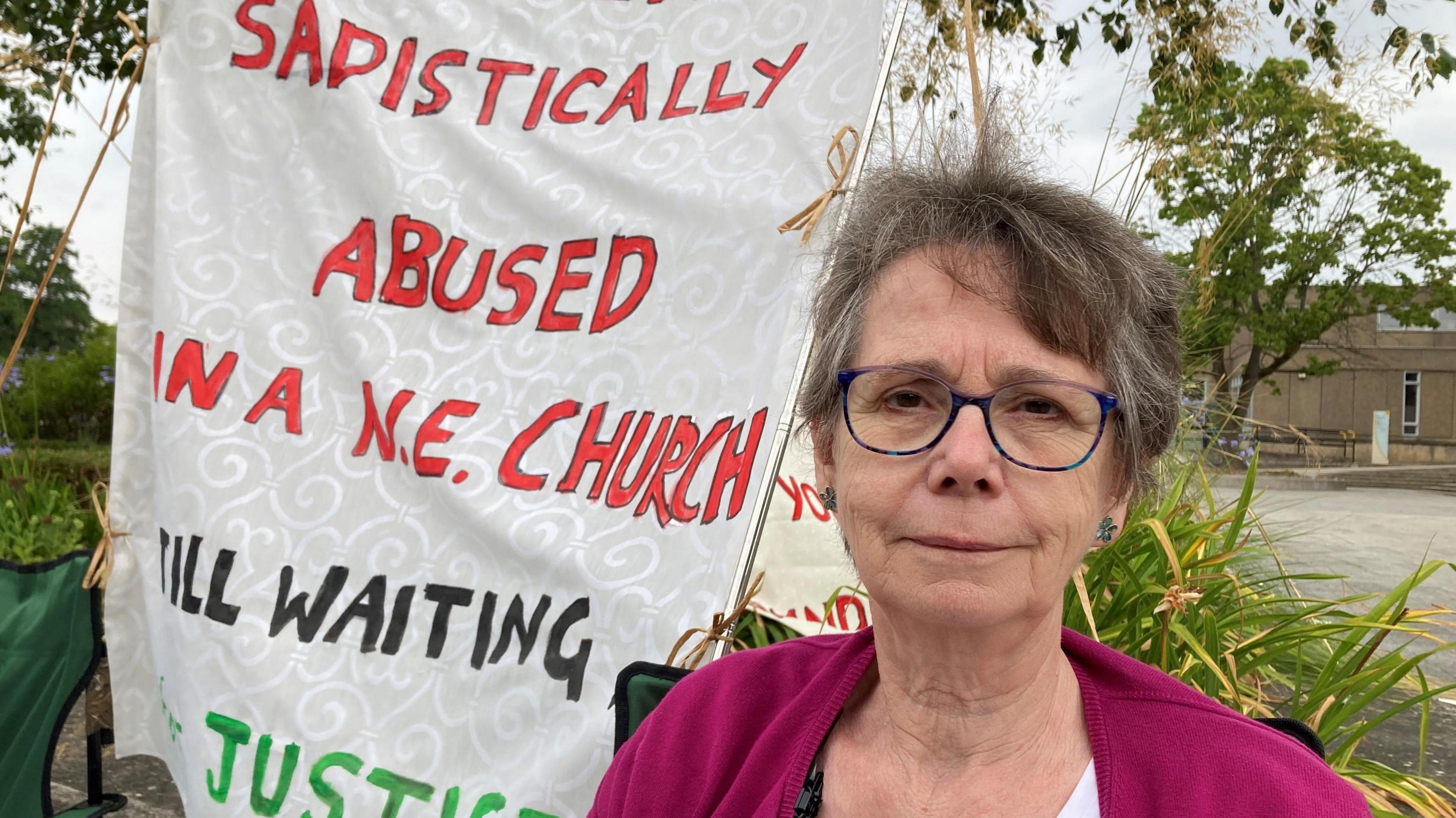 Jenny Read is sitting on a camping chair protesting outside the Church of England's parliament, the General Synod. Behind her is a banner which reads 'sadistically abused in a NE church, still waiting for justice' - although the word "for" is hidden in the fold of the banner. It's written in red, green and black on what looks like embossed, cream furnishing or curtain fabric.