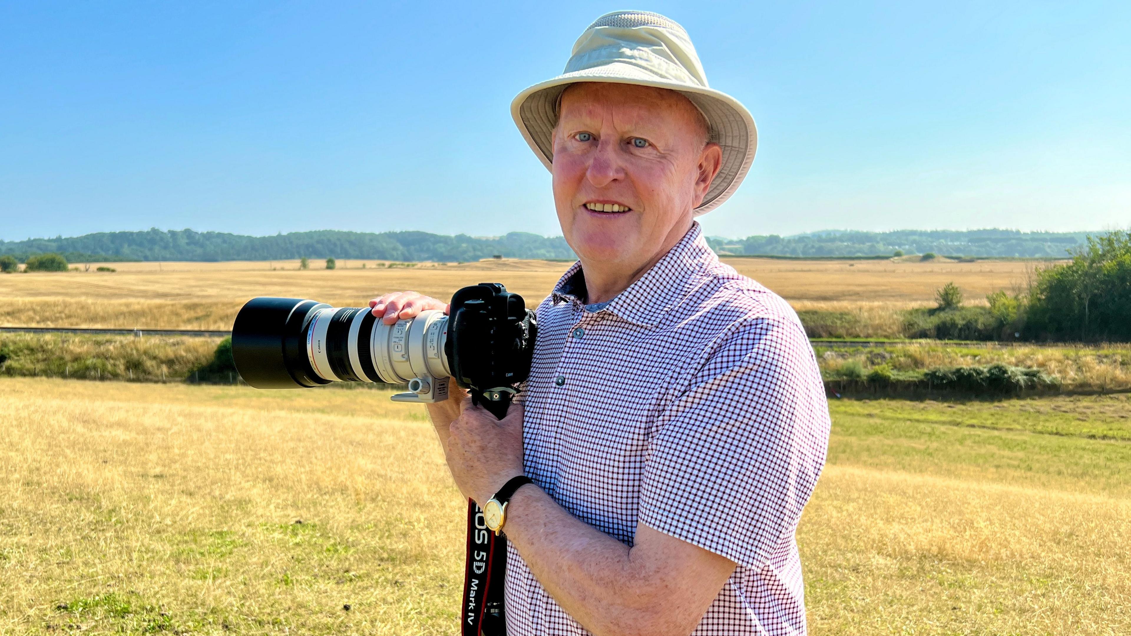 A man in a check, short sleeved shirt and cream sun hat stood in a field overlooking a railway line with a camera and tripod