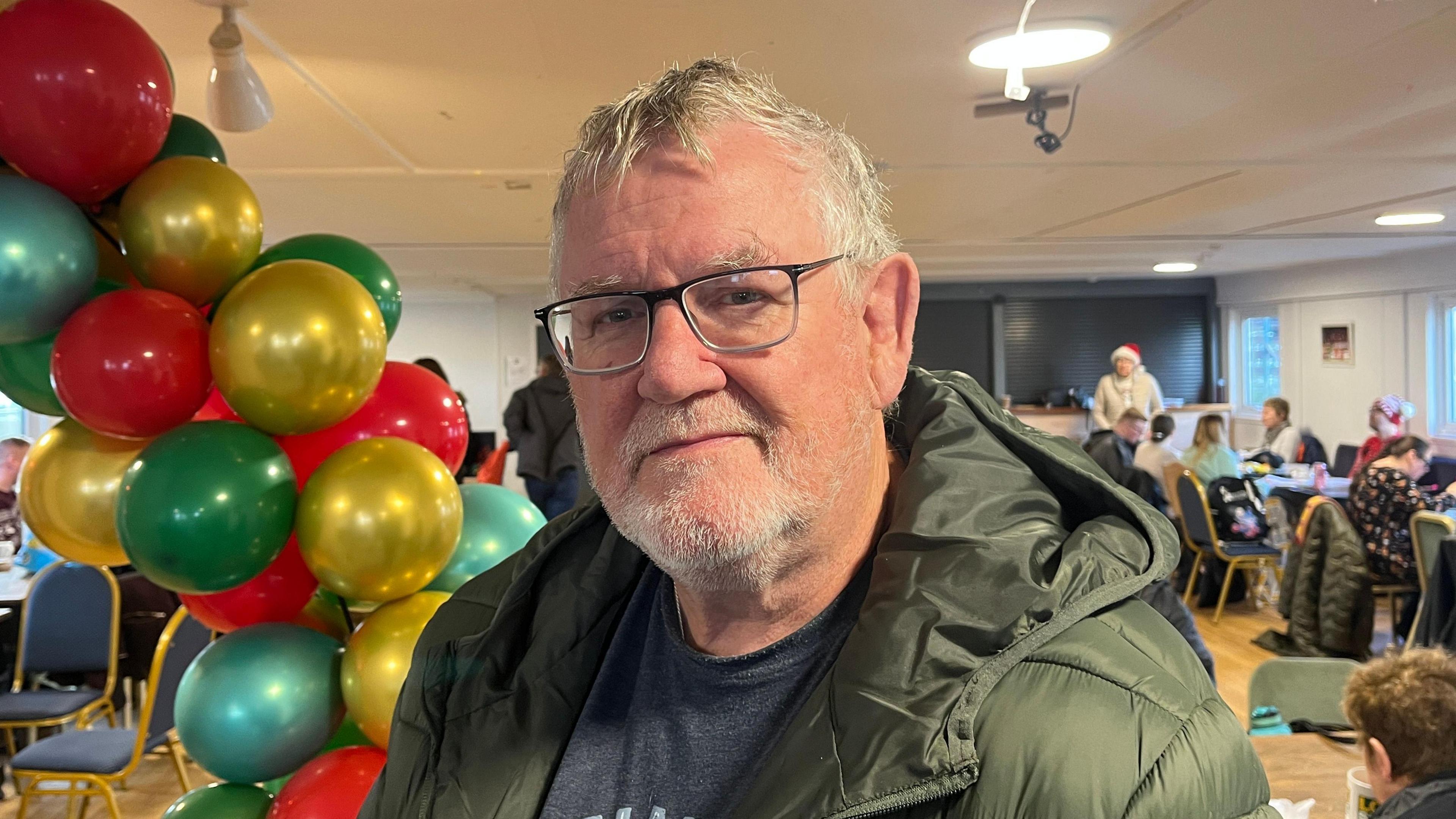 Dyn yn gwisgo sbectol â barf fer yn edrych yn syth ymlaen. Mae ganddo got werdd ac mae'n sefyll mewn ystafell gyda phobl yn eistedd y tu ôl iddo. Mae balwns amryliw hefyd y tu cefn iddo.