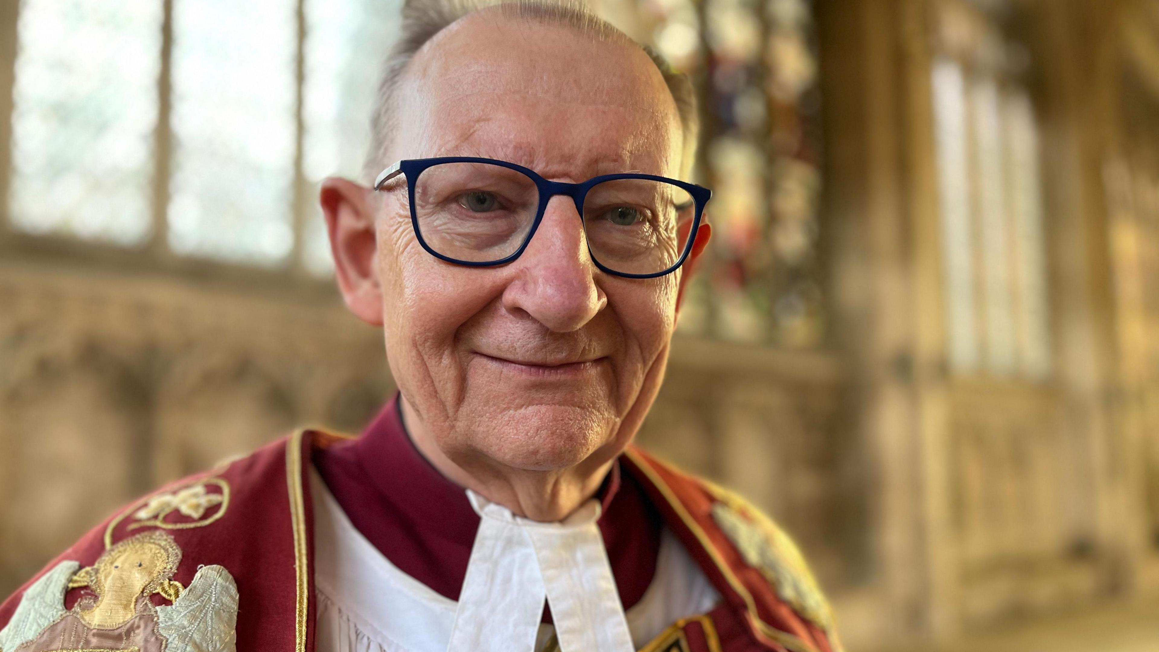 Chris Dalliston looks directly at the camera as he is photographed in Peterborough Cathedral wearing a red ceremonial cloak over his red dress. Behind him is a number of stained-glass windows. He is wearing blue-rimmed glasses. 