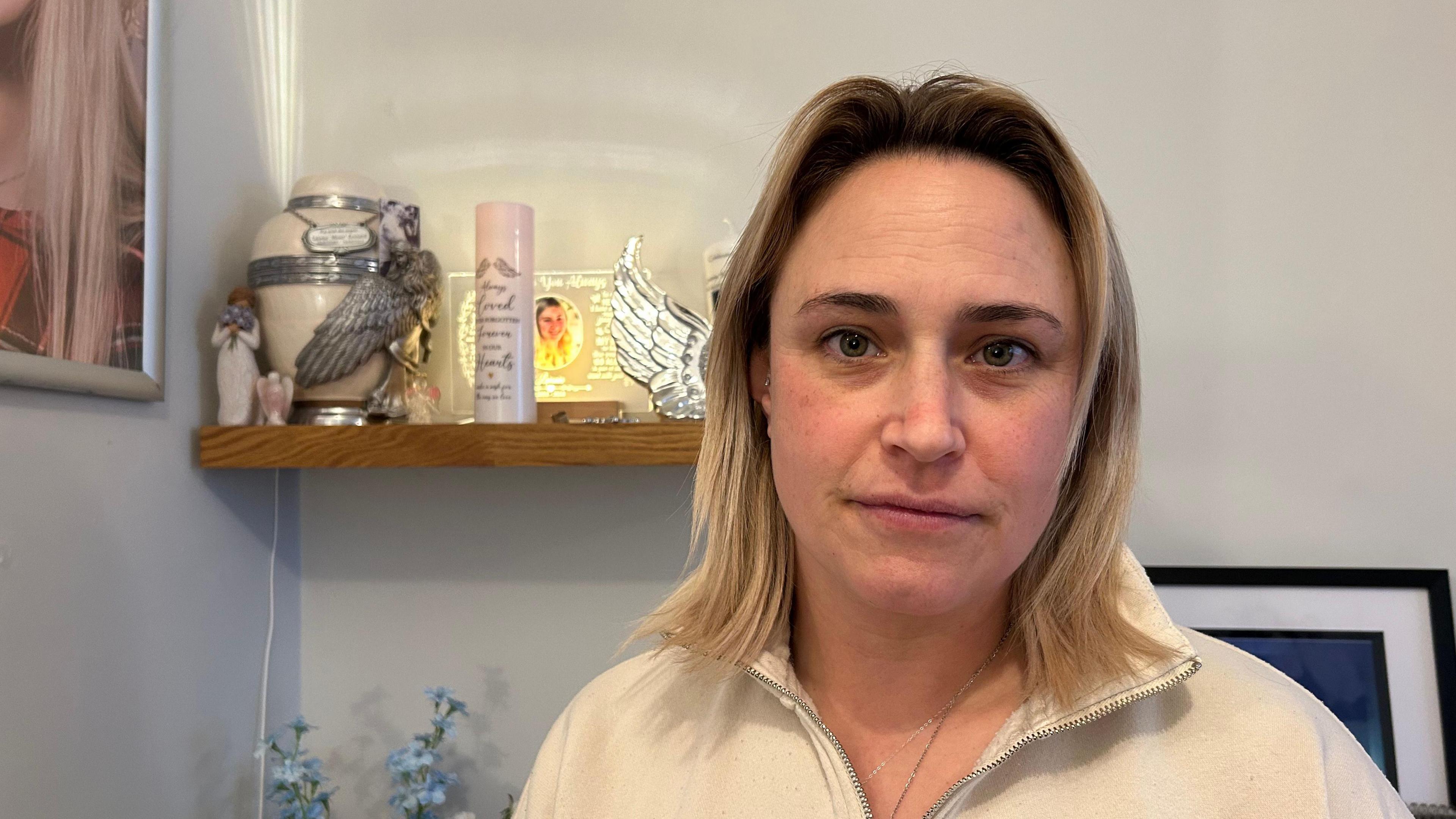 A woman wearing a beige fleece - behind her is an urn of ashes, a candle and a picture of her daughter. 