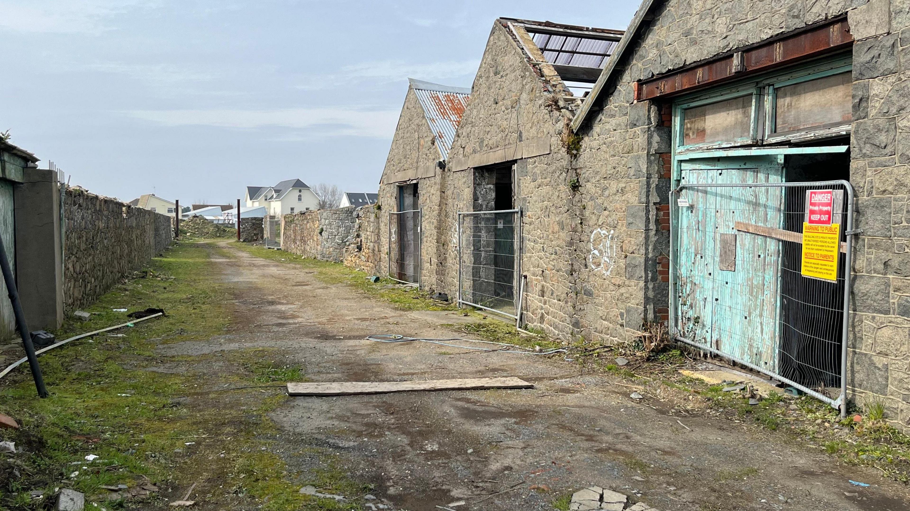 A number of derelict brick buildings with fences and signs on their doors - one of which says 'danger keep out'. The ground has bricks and moss on it. 