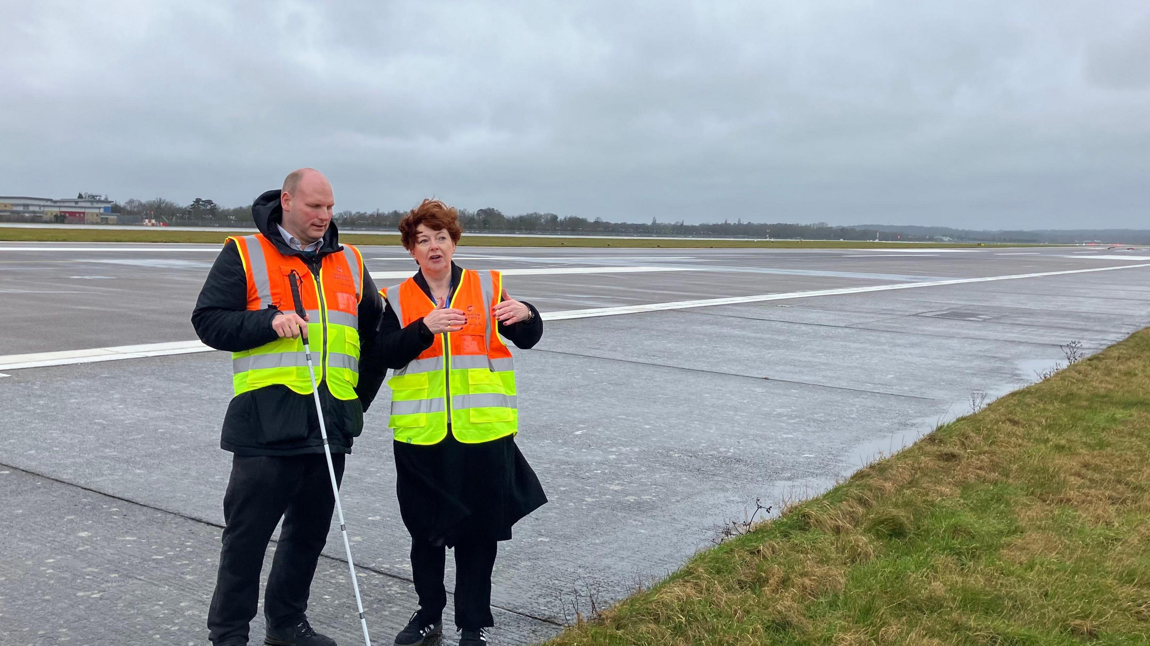 Bronwen Jones, Development Director at London Gatwick standing on the northern runway at Gatwick airport alongside BBC transport correspondent Sean Dilley, pointing to a grass verge where the runway will be expanded under the airport's proposals. Both are wearing high vis jackets.