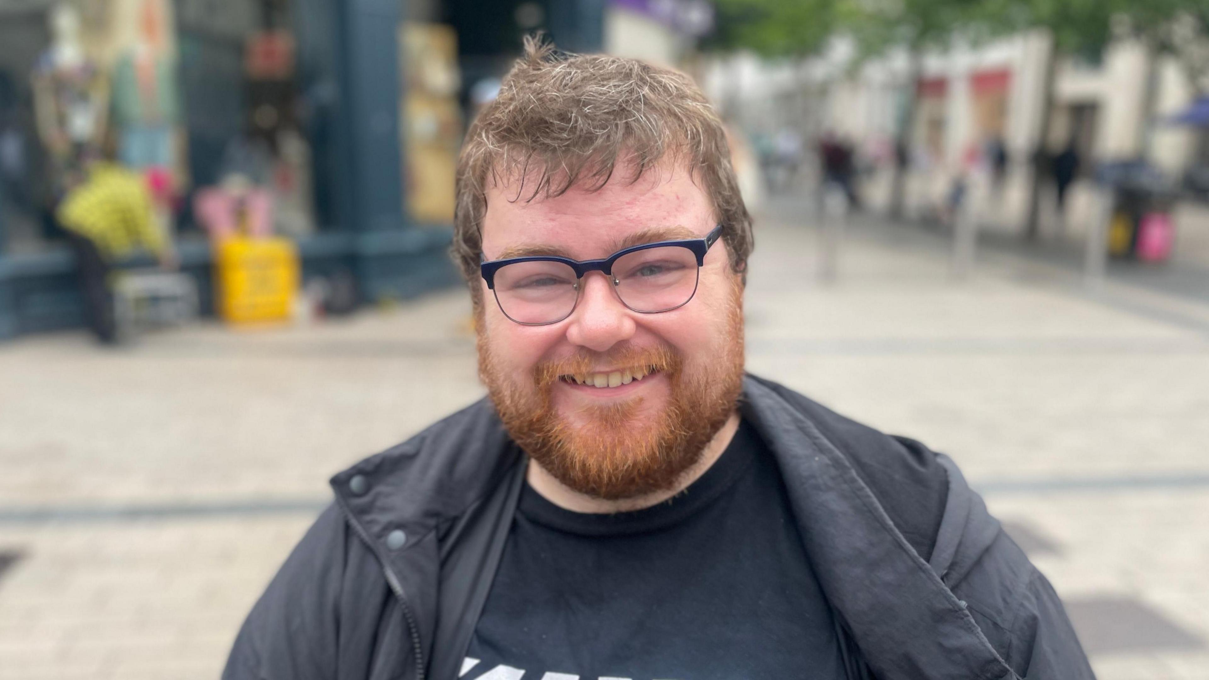 Paul Maye is wearing a black coat and black t-shirt, he has black glasses on. He has ginger facial hair and brown head hair. The background is blurry. 