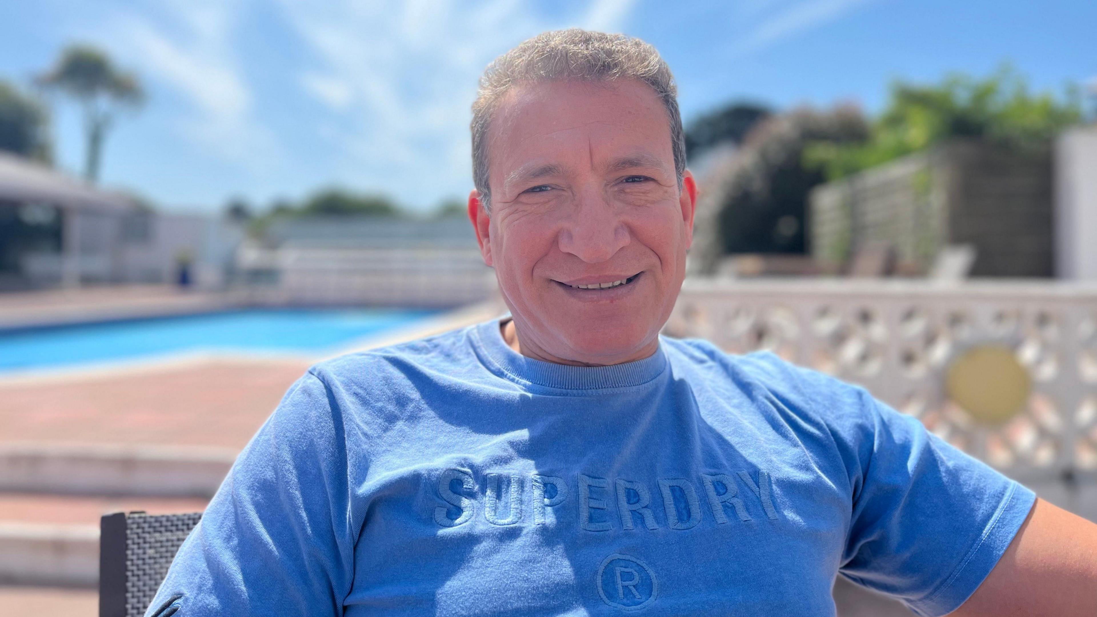 Photo of a man with short brown hair, smiling at the camera. He is wearing a light blue t-shirt and is sat on a chair with a swimming pool in the background