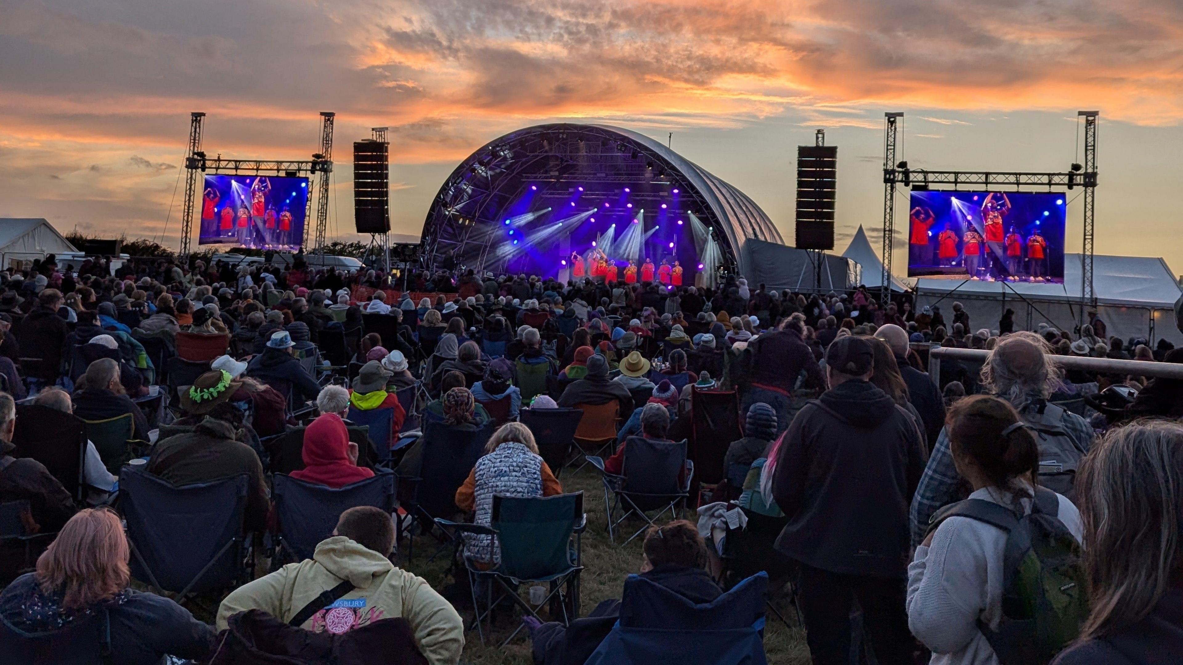 A crowd watch on at Shrewsbury Folk Festival. A large semi-circle shaped stage with screens either side of it is seen in the shot.